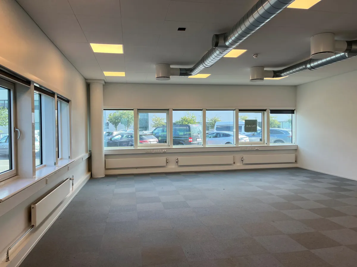 Kontor med møbleret gulv og radiatorer. Store ruder med udsigt til parkeringsplads med biler og bus. Loft med ventiler og