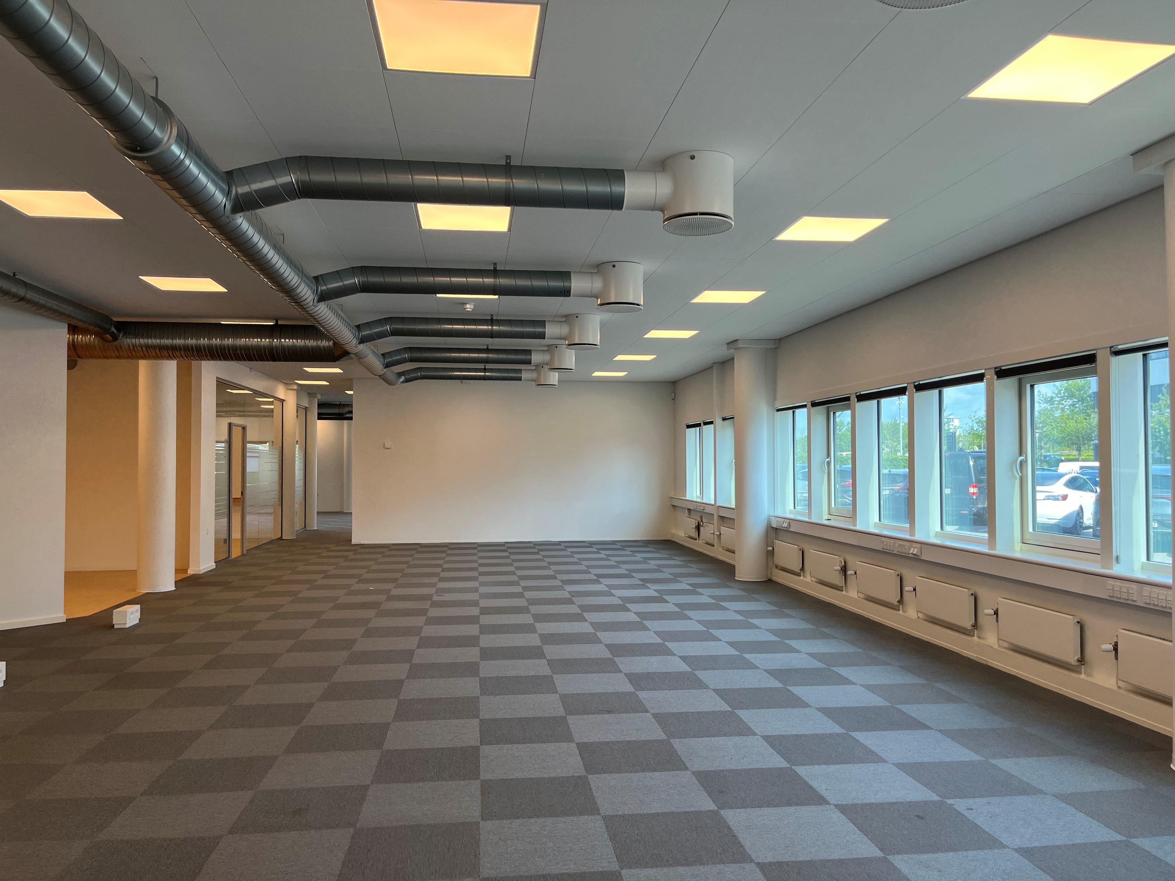 Værelse med krydsfinerbelagt gulv, hvide vægge, store vinduer og slanke stålrør under loftet