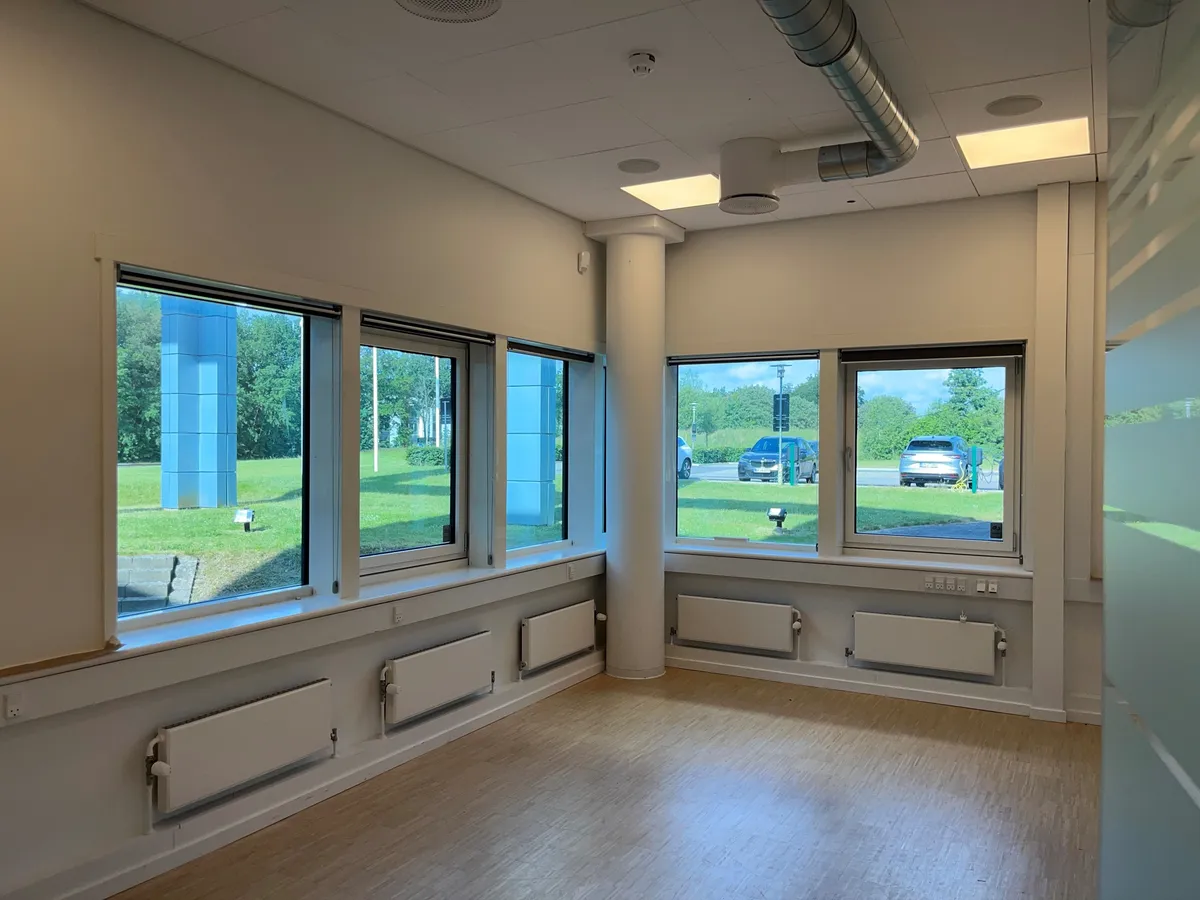 En tomt kontor med vinduer og radiatorer i hvid farve og trægulv med udsigt til grønne områder og biler parkeret på siden af