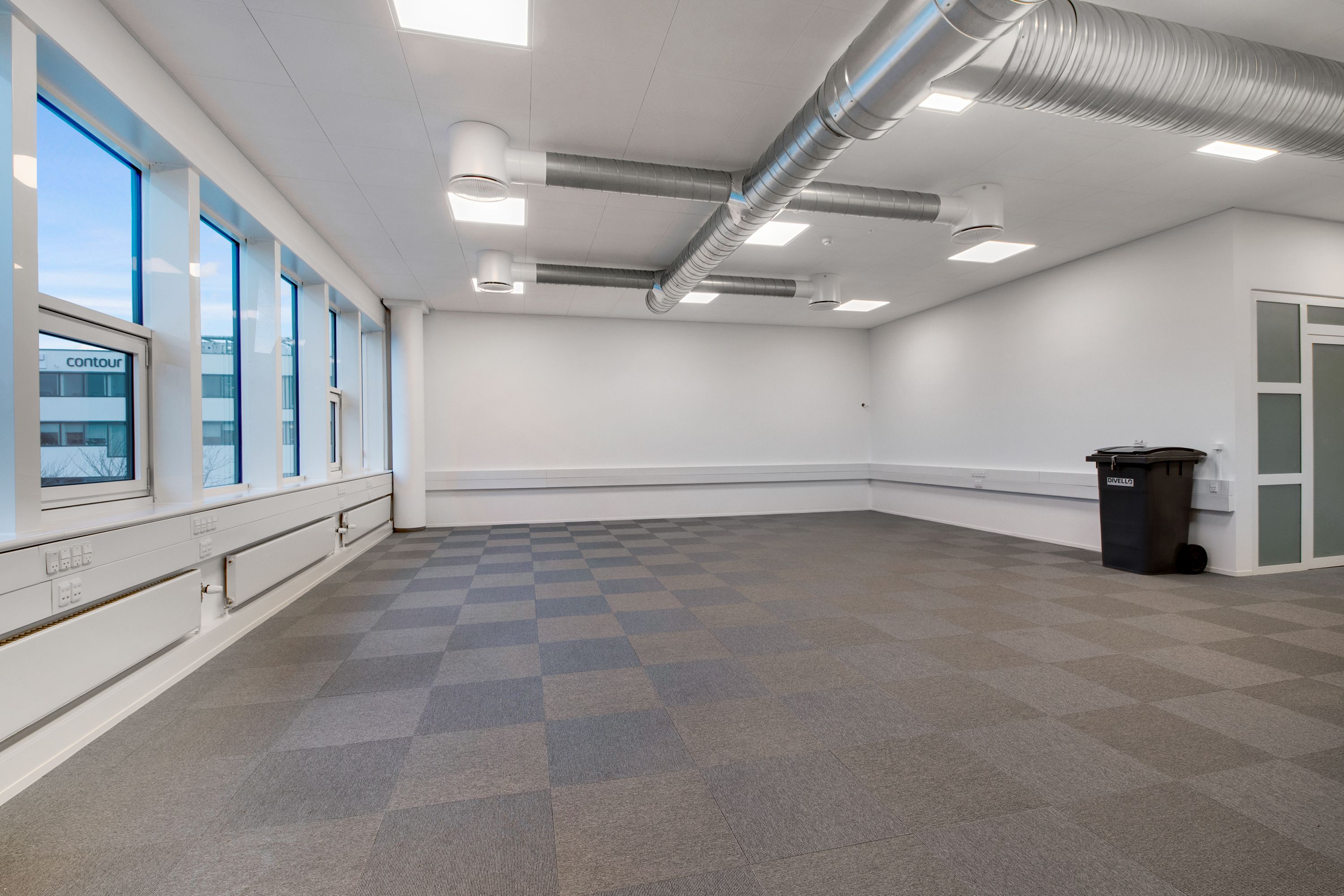 Tomt kontor med karreleret gulv og vinduer med udsigt til bygning. Hvide vægge, gulv og loft med ventilationsrør og