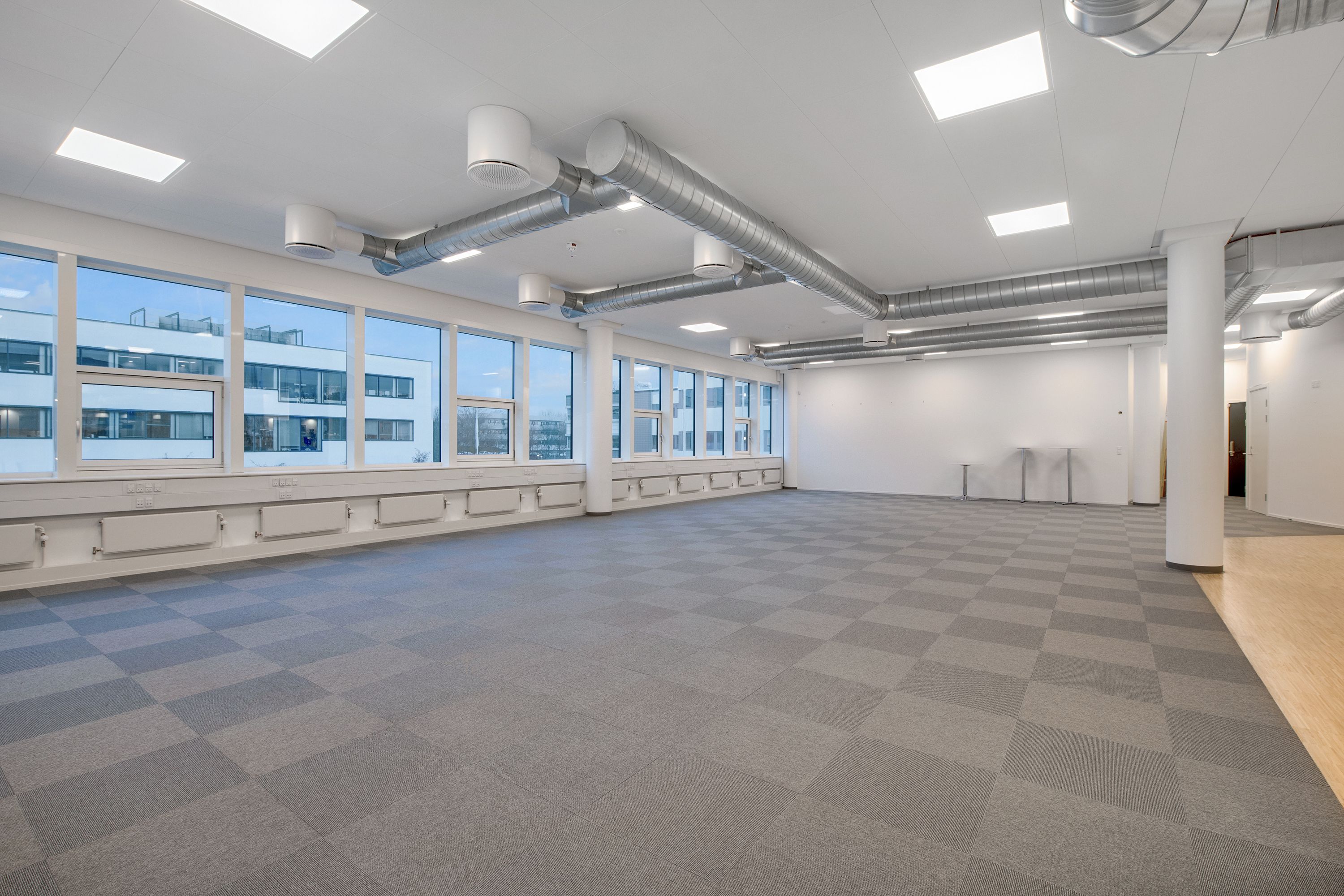 Røgsugtt med ventiler og gulvbelægning i en kontorfællesskab med vinduer mod bygningen udenfor og lyse loftslamper