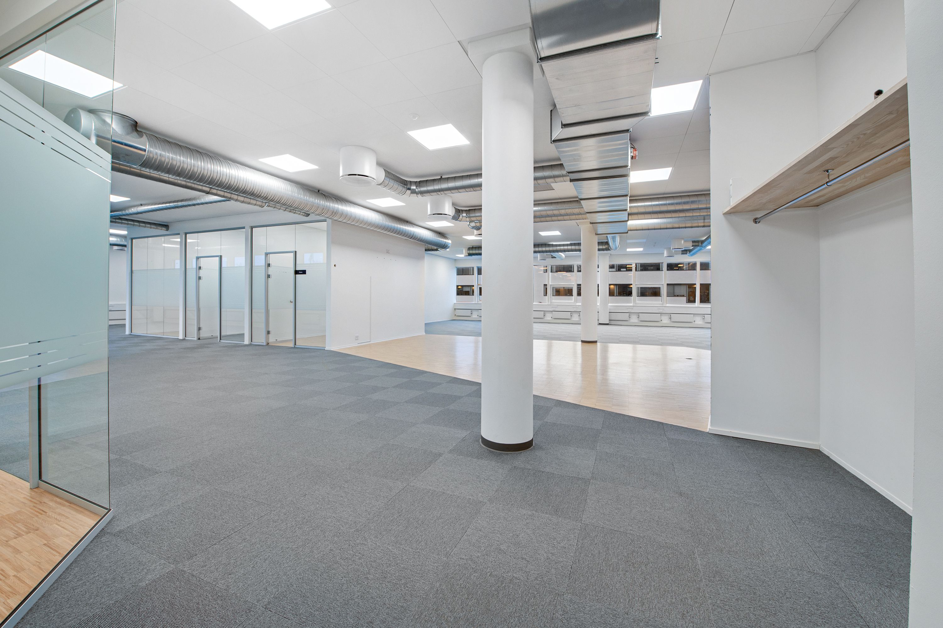 Moderne kontor med højloft og lyse loftsbjælker, hvide vægge, glasparti og gulvtæppe. Et søjle og en åben dør til et lager