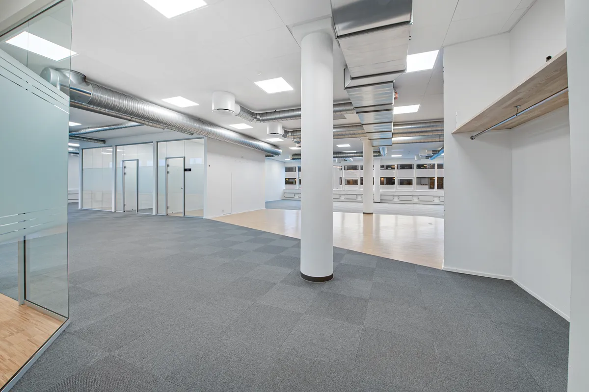 Moderne kontor med højloft og lyse loftsbjælker, hvide vægge, glasparti og gulvtæppe. Et søjle og en åben dør til et lager