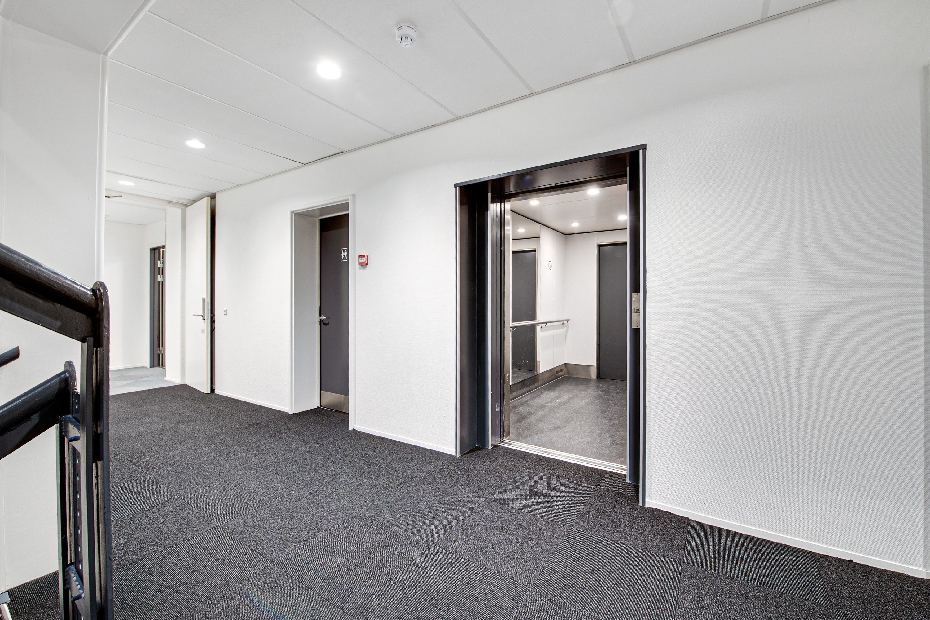 Elevator med åben dør og lysegult gulv, slank trappe med sort metal, hvide vægge og lysekroner i loftet