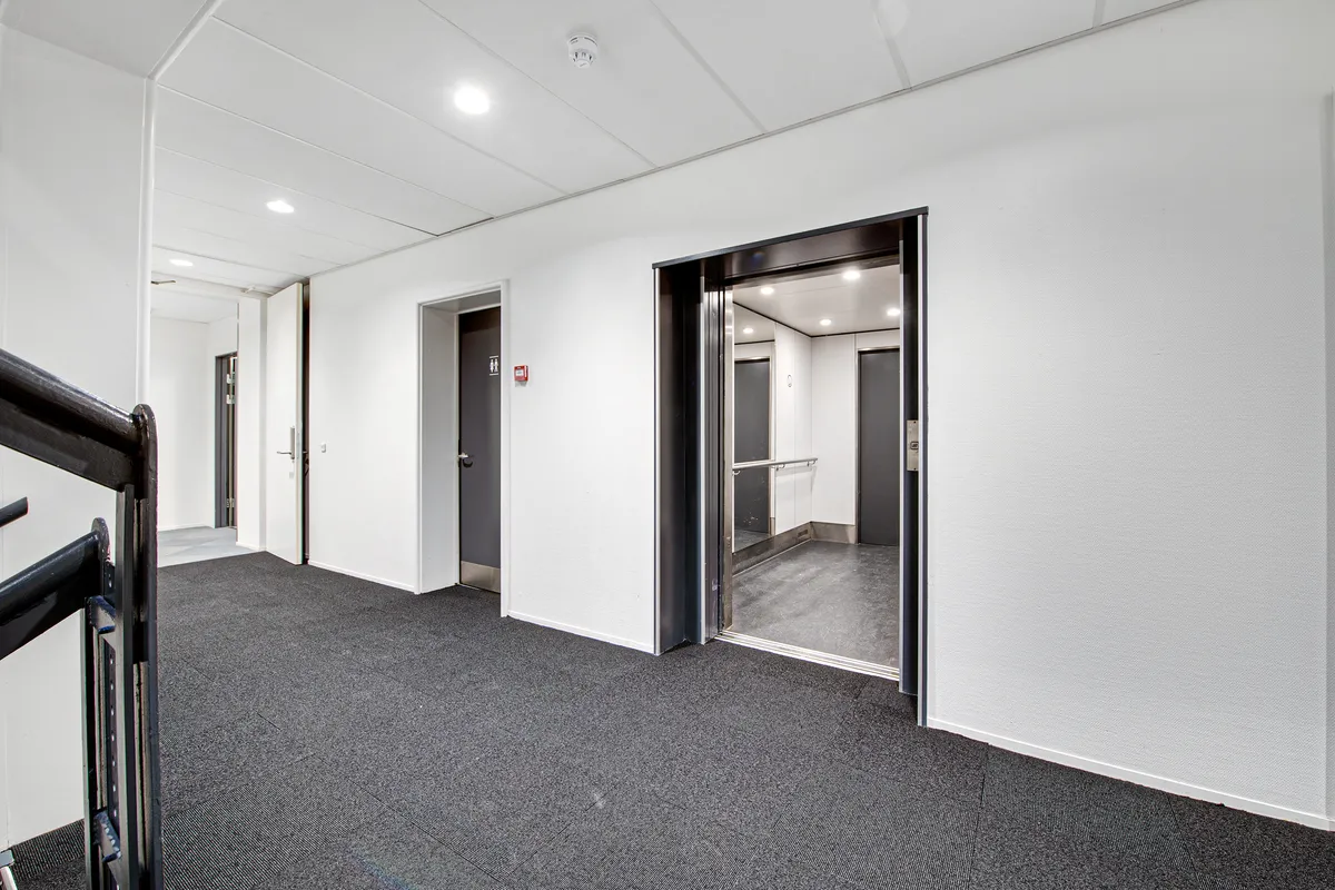 Elevator med åben dør og lysegult gulv, slank trappe med sort metal, hvide vægge og lysekroner i loftet