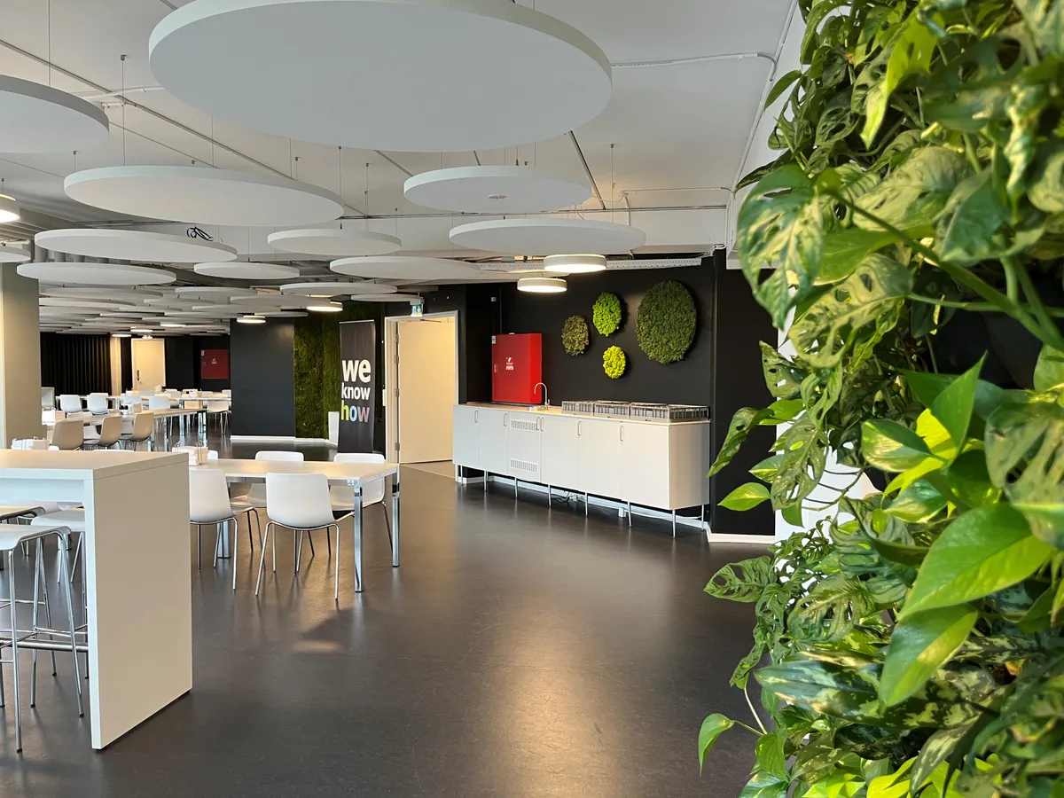 Kontor med rundt loft, plantevæg, rødt køleskab, lyse høje stole og hvide spisesteder med runde borde og lyse bænke.