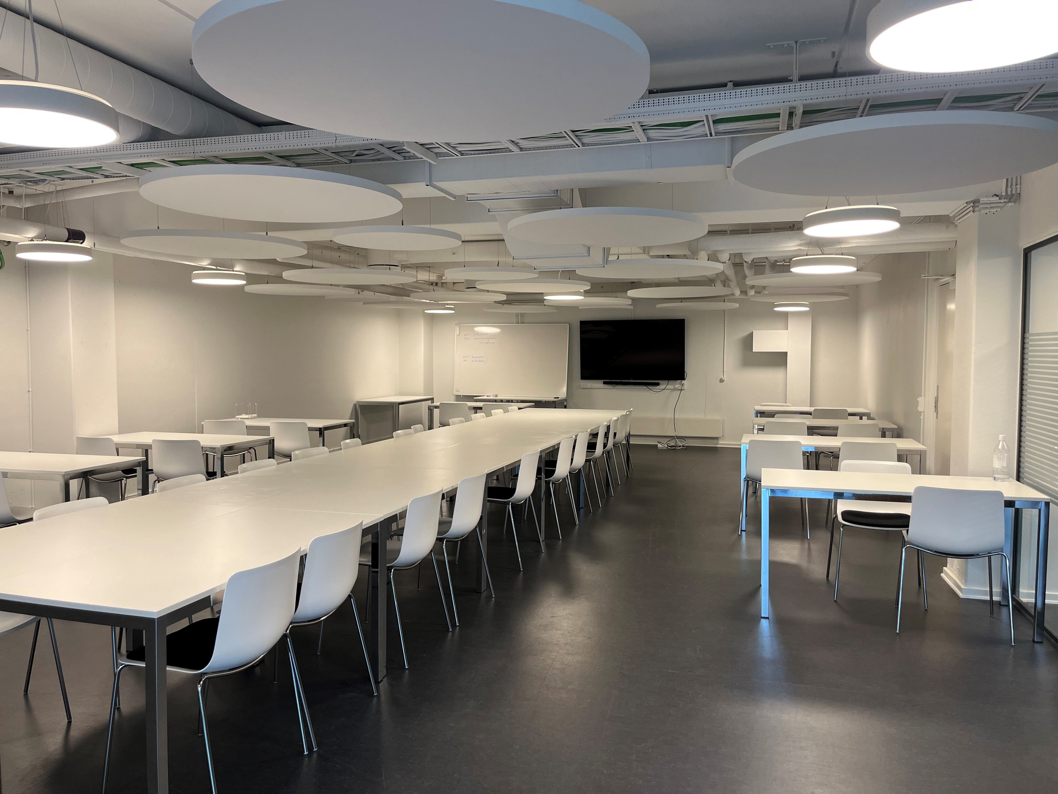 Kontor med lyse loftslys og runde lysekroner, flere lange borde med hvide stole og tv og whiteboard på væggen