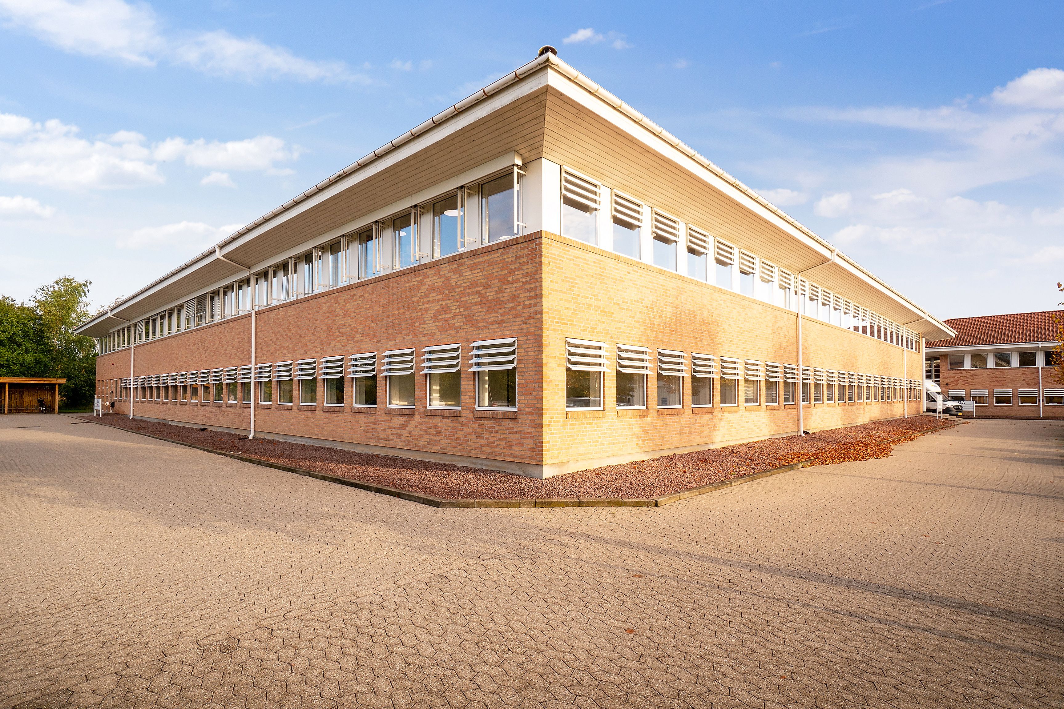 Kontorbyggeri med flere vinduer og garage på en stor parkeringsplads under en blå himmel med nogle skyer