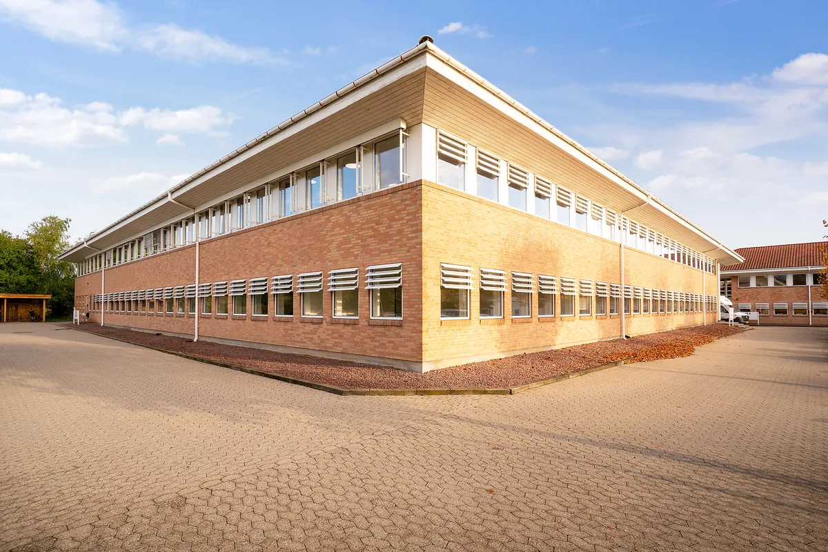 Kontorbyggeri med flere vinduer og garage på en stor parkeringsplads under en blå himmel med nogle skyer
