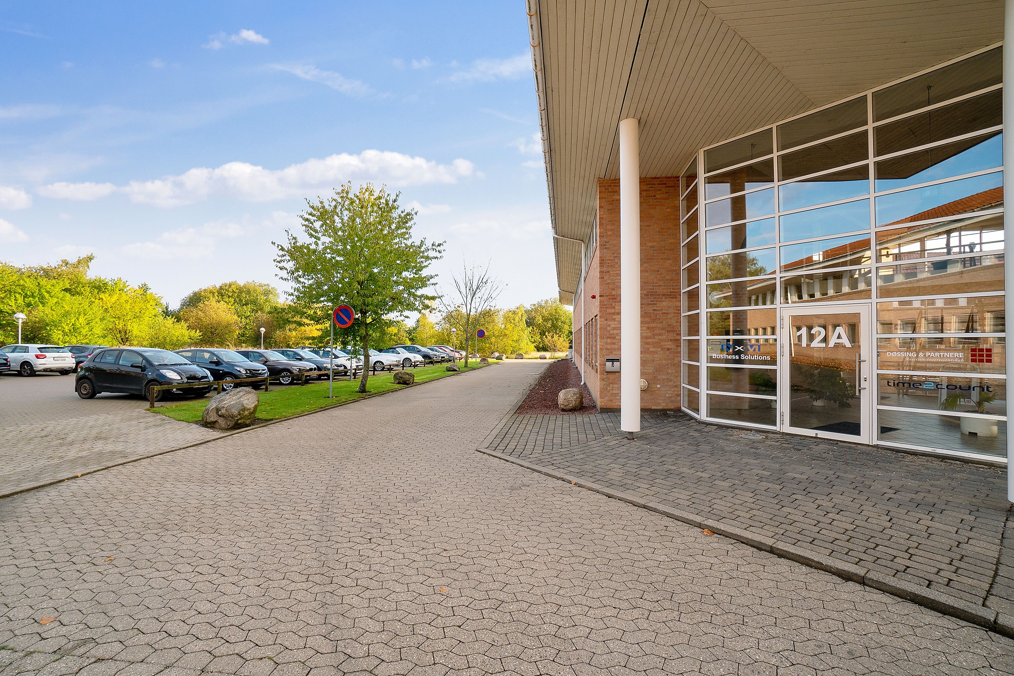 Kontorbygning med moderne design, glasvinduer og parkeringsplads for biler. Der er en gågade foran bygningen og en række
