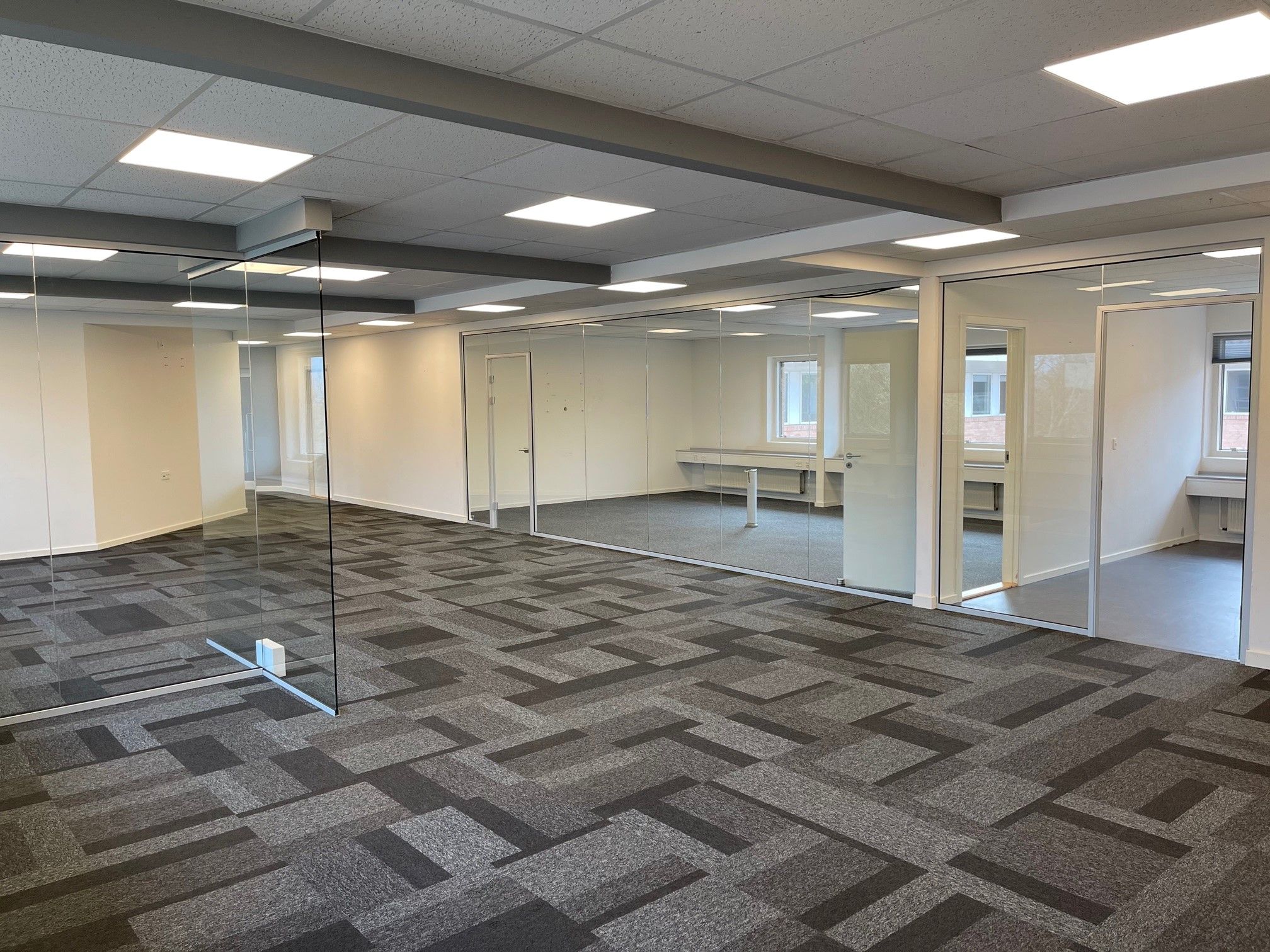 En stor, møbleret kontor med store glasvægge og et lysegråt loft med indlejrede lys. Der er et stort åbent område med