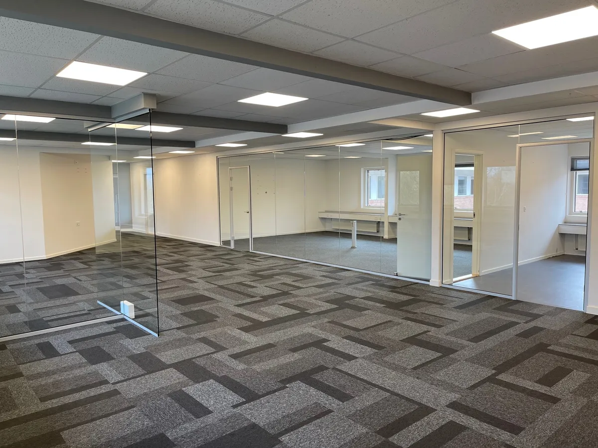 En stor, møbleret kontor med store glasvægge og et lysegråt loft med indlejrede lys. Der er et stort åbent område med