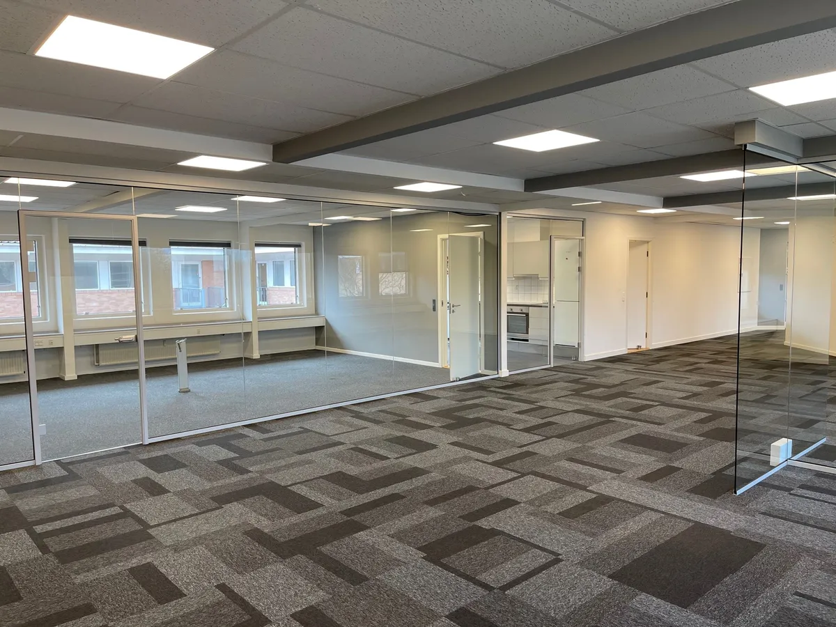 Et moderne kontor med glasvægge og lysegulv samt hvide vægge og lyse loftsbelysninger. Et kabinet med hvide skabe og en hvid