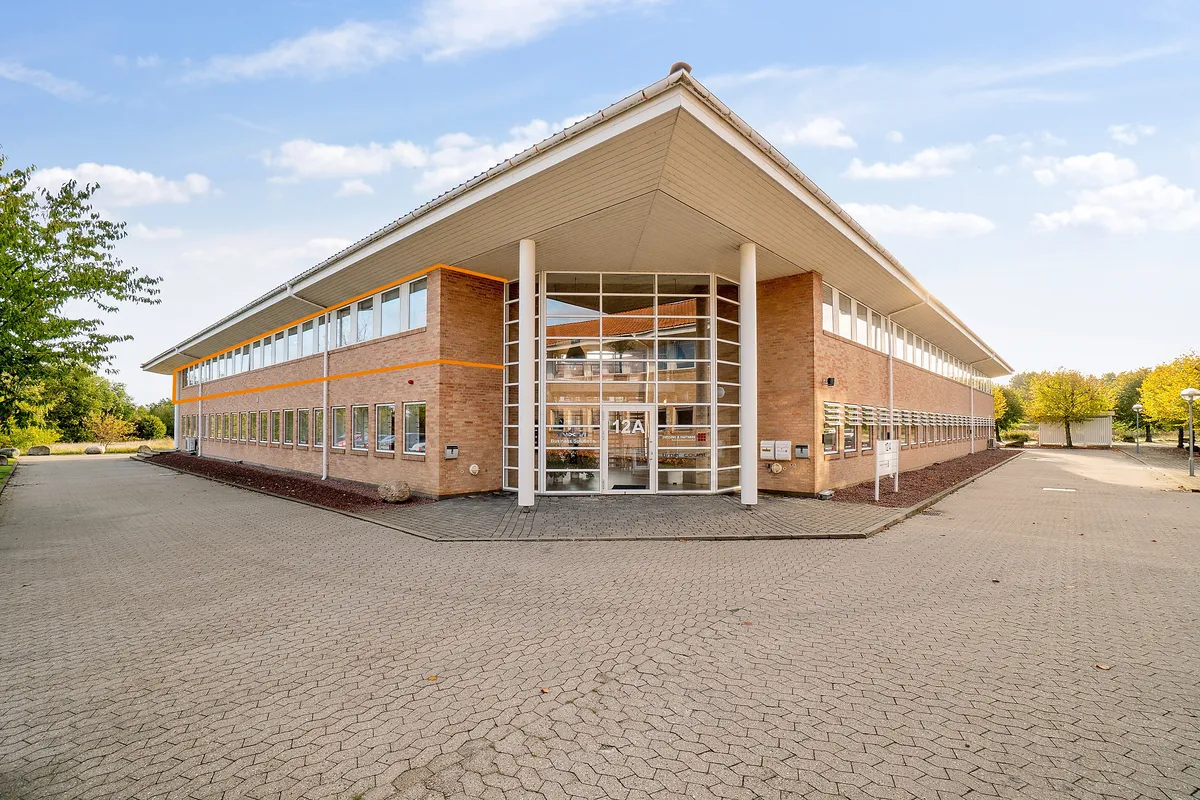 Kontorby i Hillerød med glasdøre og vinduer i orange og hvidt udsmykning. Parkeringsplads foran bygningen og et slankt træ i