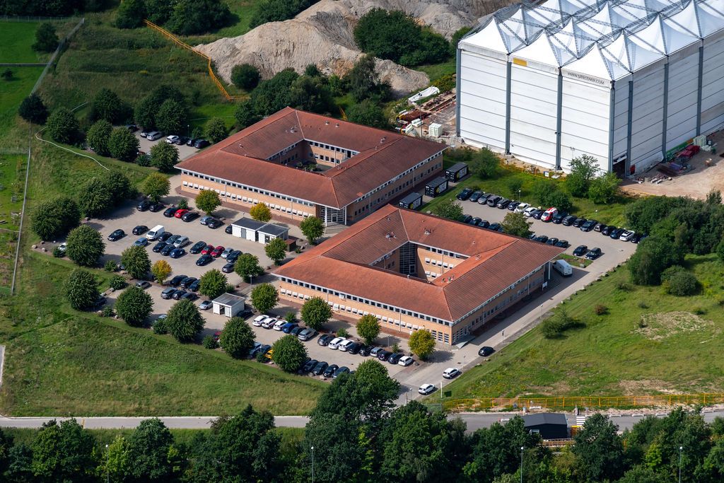 Bygninger med parkeringspladser for biler i området med grønne træer og buske. Et stort lager og jordhøje ligger i