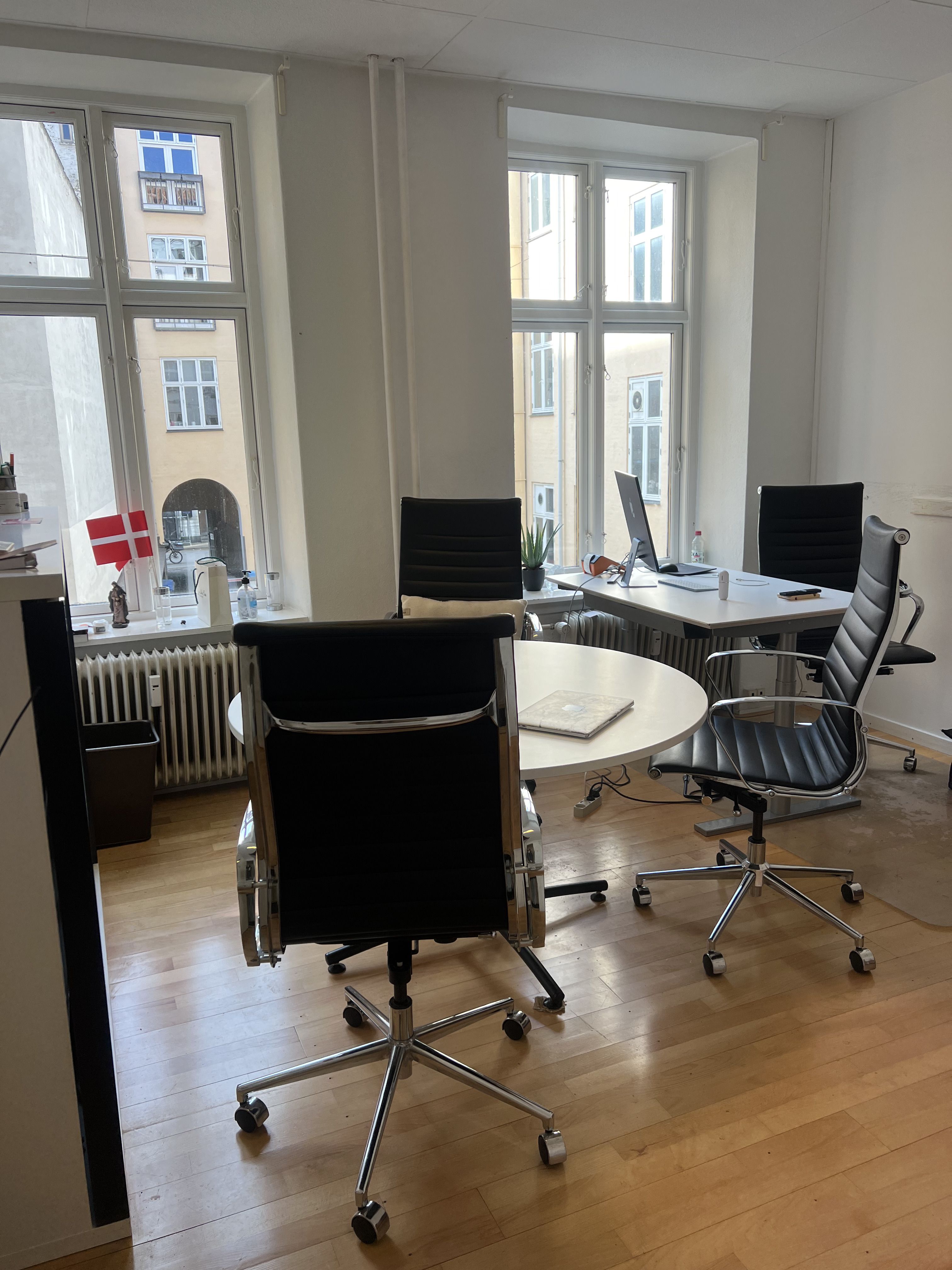Kontor med rundt mødebord og fire læderstole, samt et skrivebord med computer og en række ting på vindueskarmen