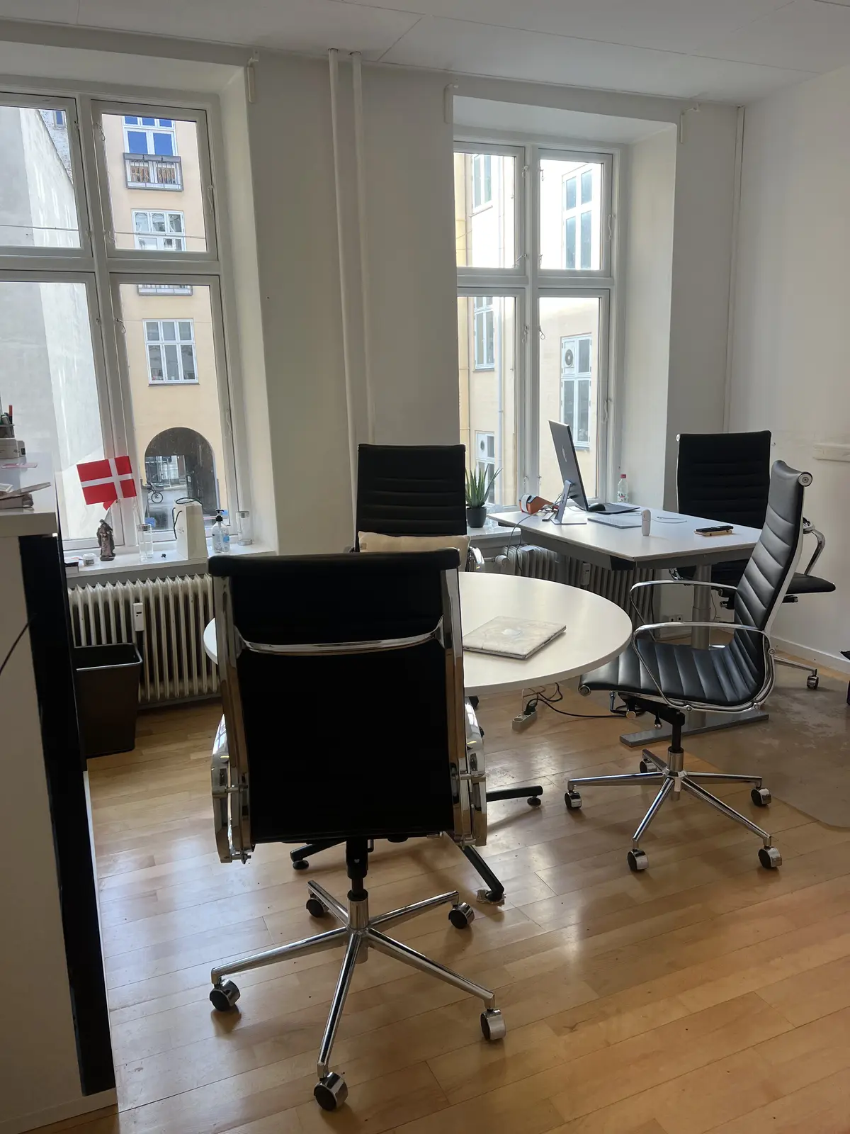 Kontor med rundt mødebord og fire læderstole, samt et skrivebord med computer og en række ting på vindueskarmen