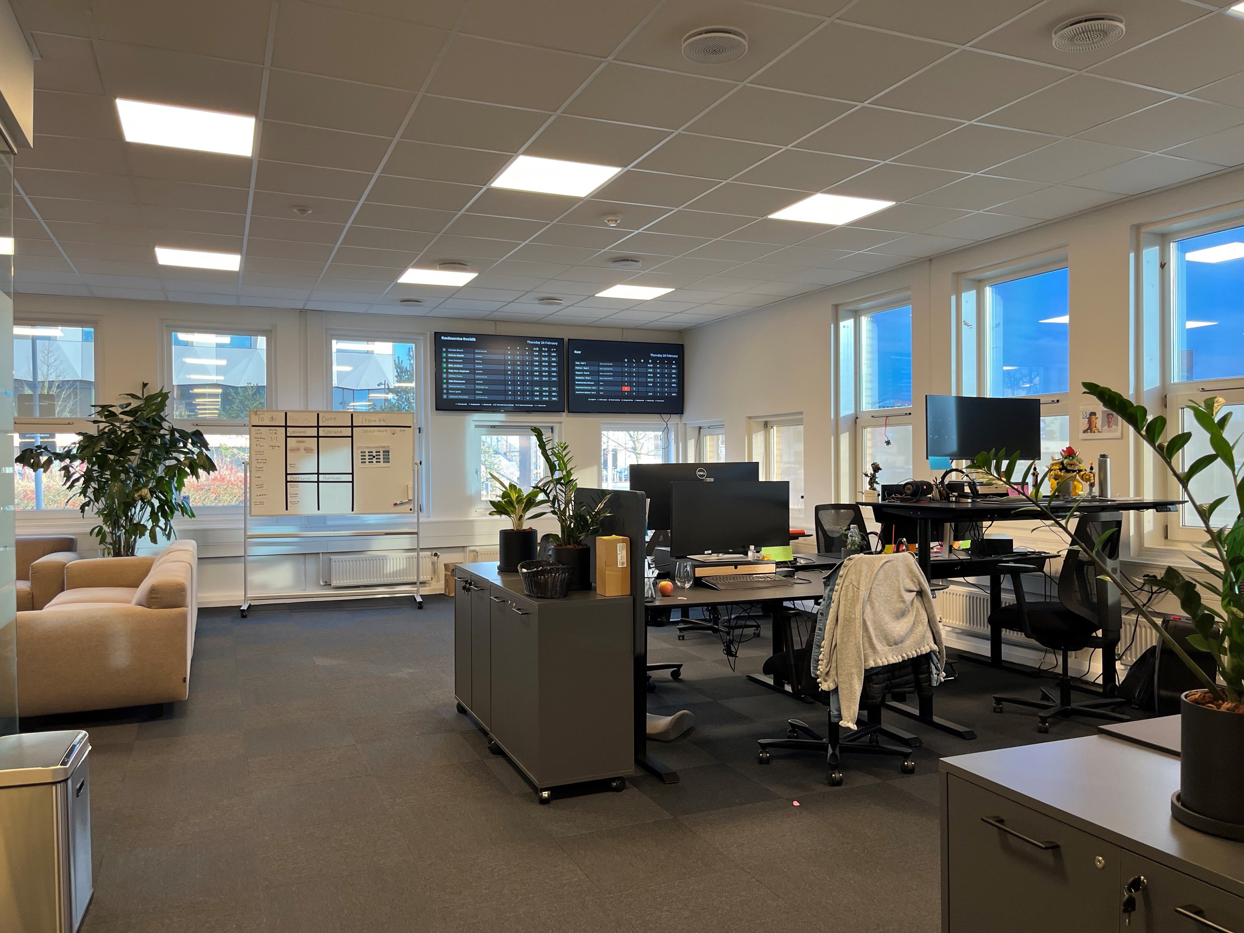 Moderne, lyse og renslige kontor med to monitorer og flere computere. Store vinduer med udsigt til udendørs og to store