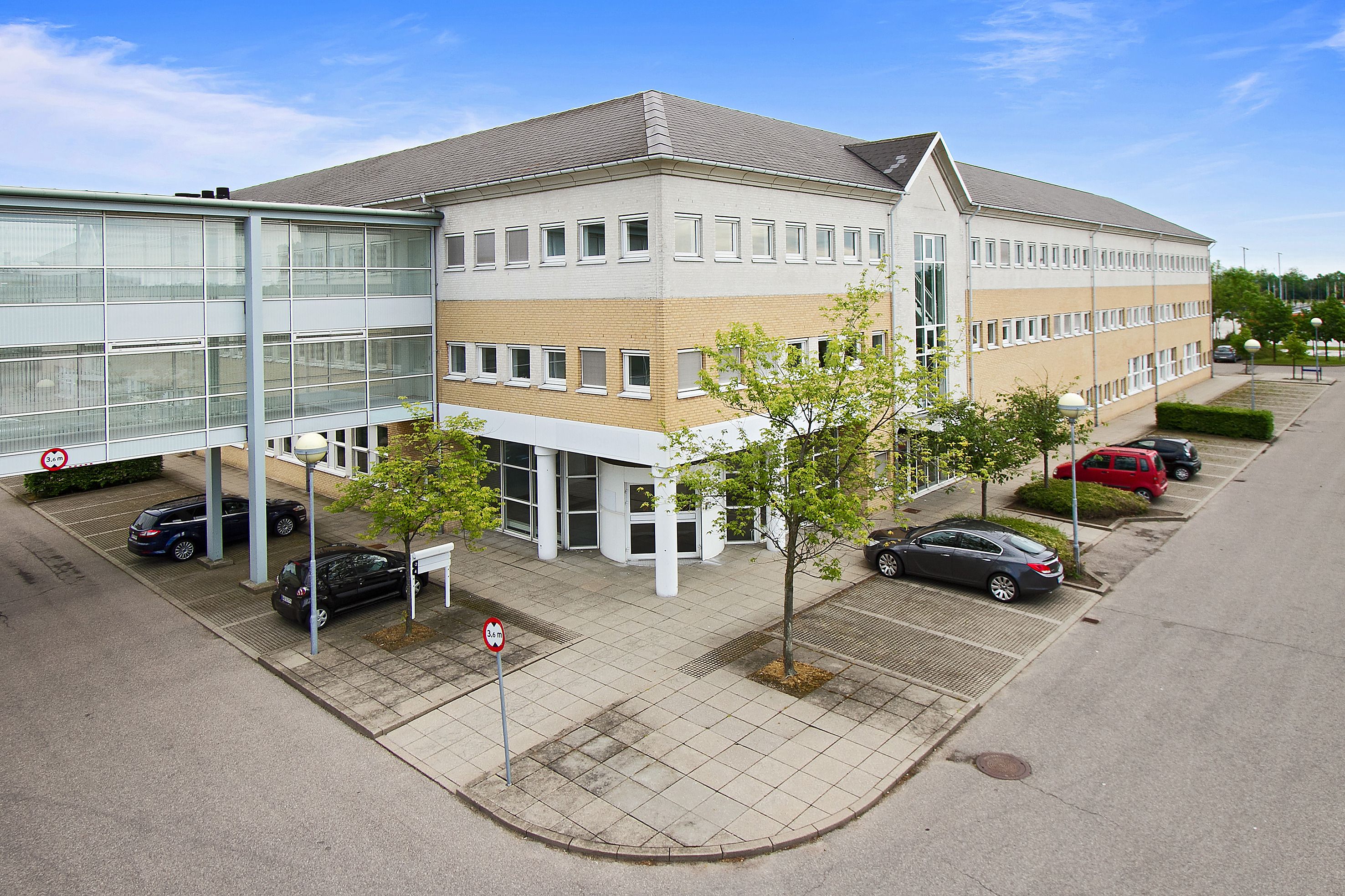 En stor bygning med flere vinduer, et glasgang og et parkeringsområde med flere biler og to vejplanlægningsskilte