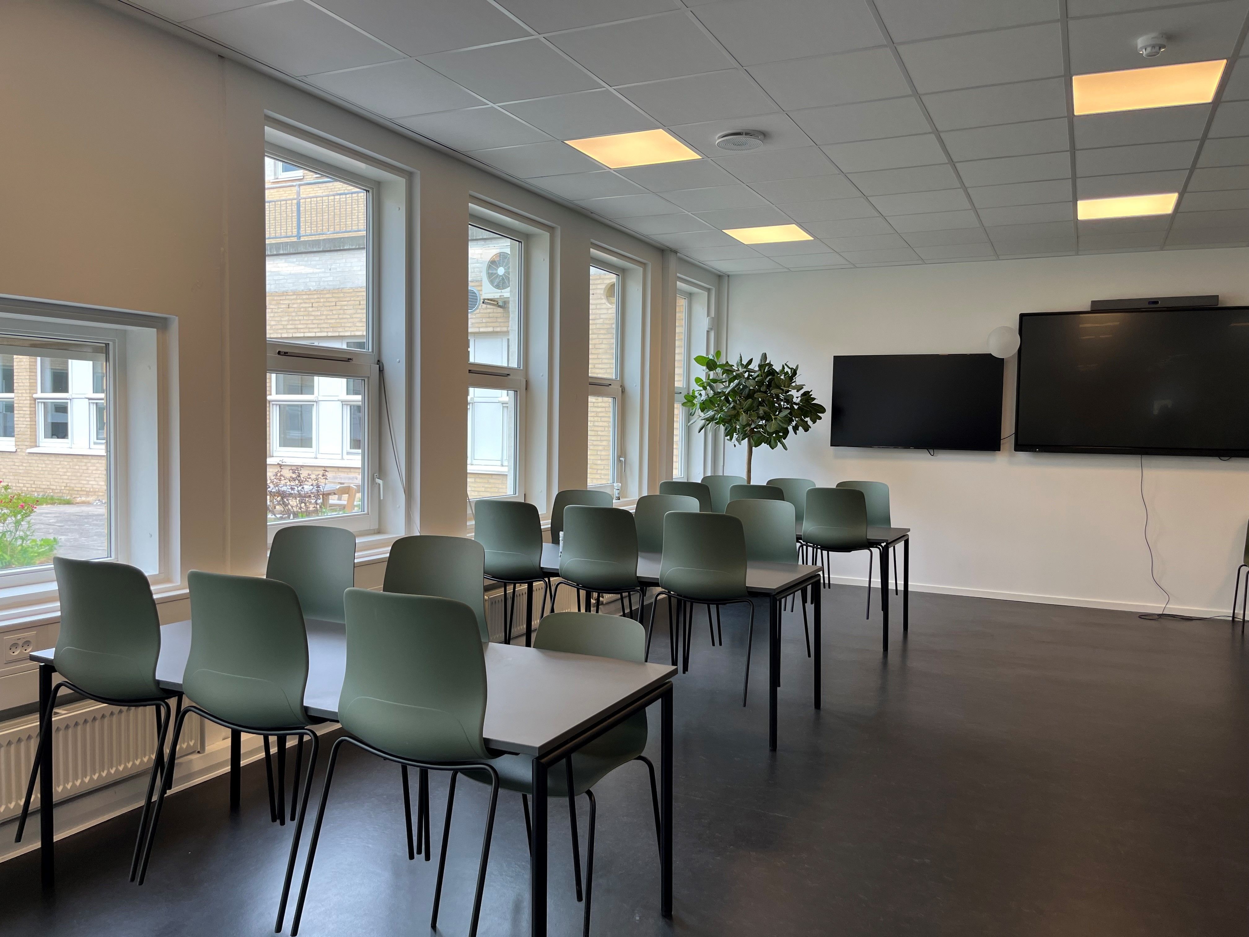 Billede af et tomt mødelokale med grønne stole og bord. To store tv-skærme er monteret på væggen. Rummet er lyst og
