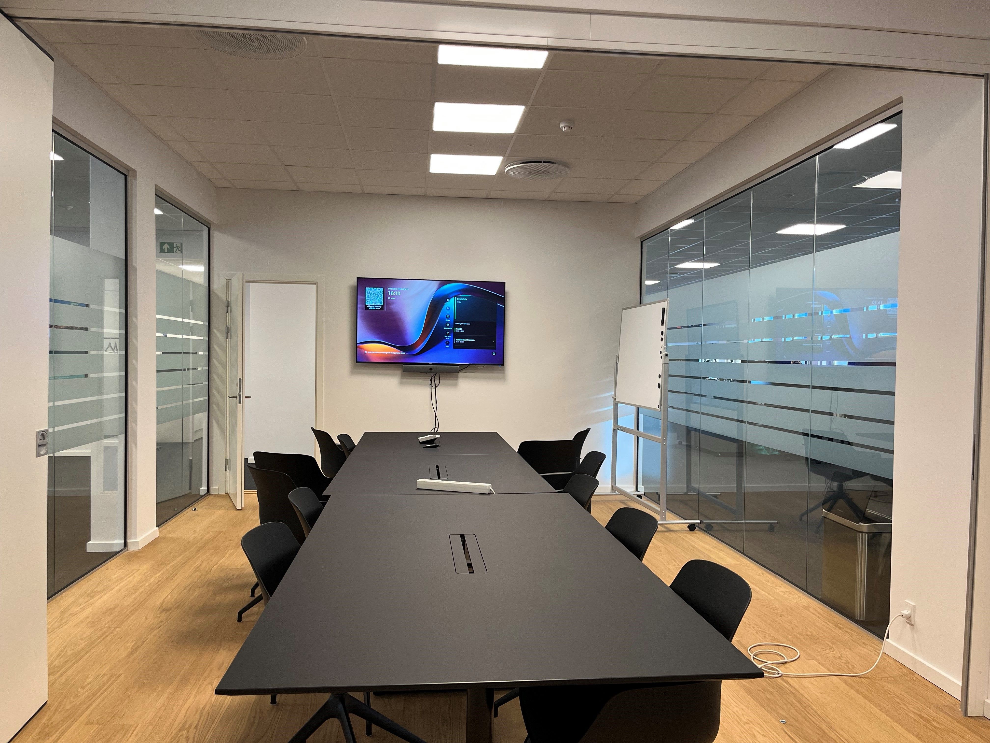 Kontorlokation med lyse loftsforhøjelser og glasvægge. Sort bord med computermus og pen. Hvidt tavle og fjernsyn. Sorte