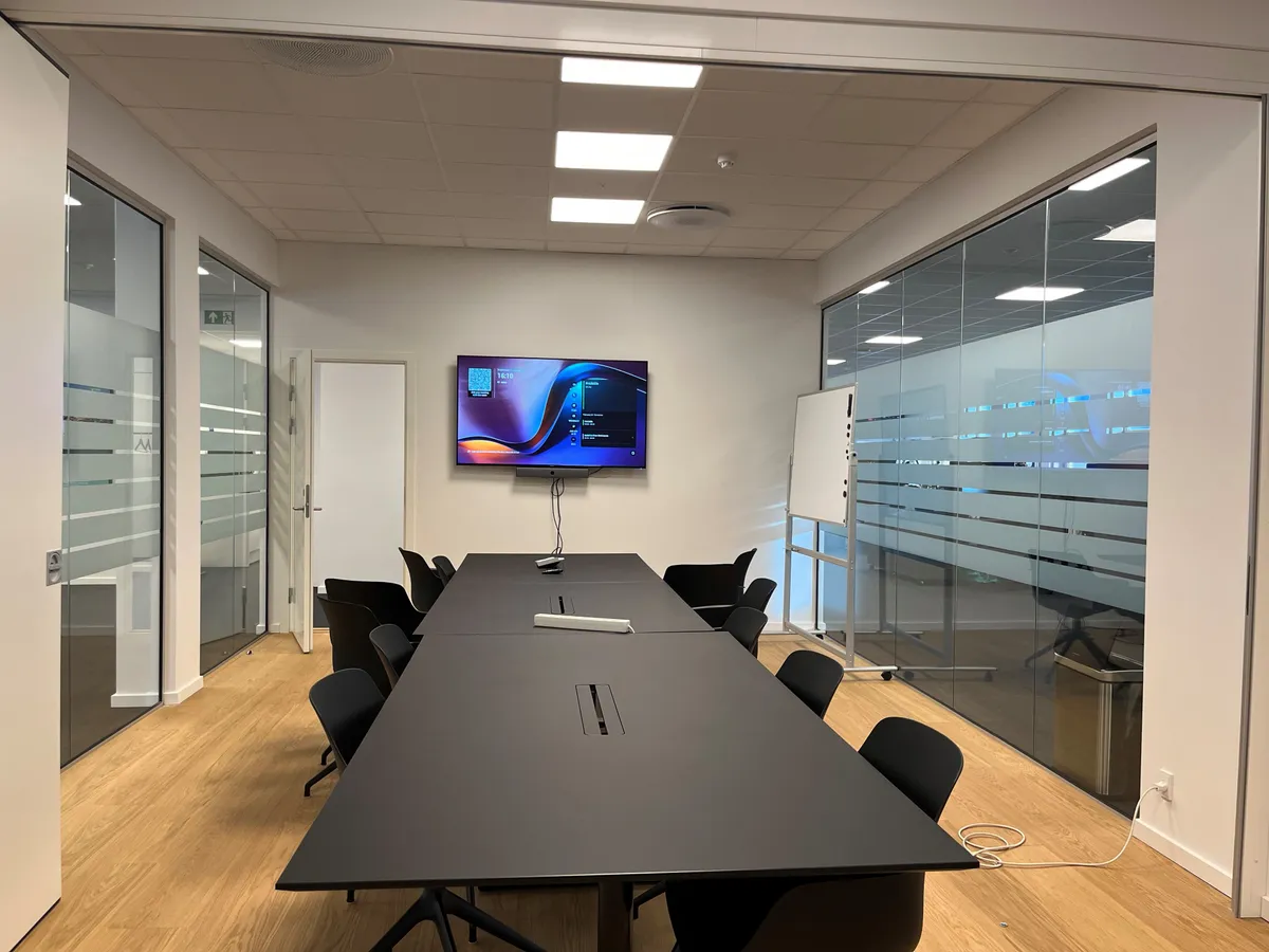 Kontorlokation med lyse loftsforhøjelser og glasvægge. Sort bord med computermus og pen. Hvidt tavle og fjernsyn. Sorte