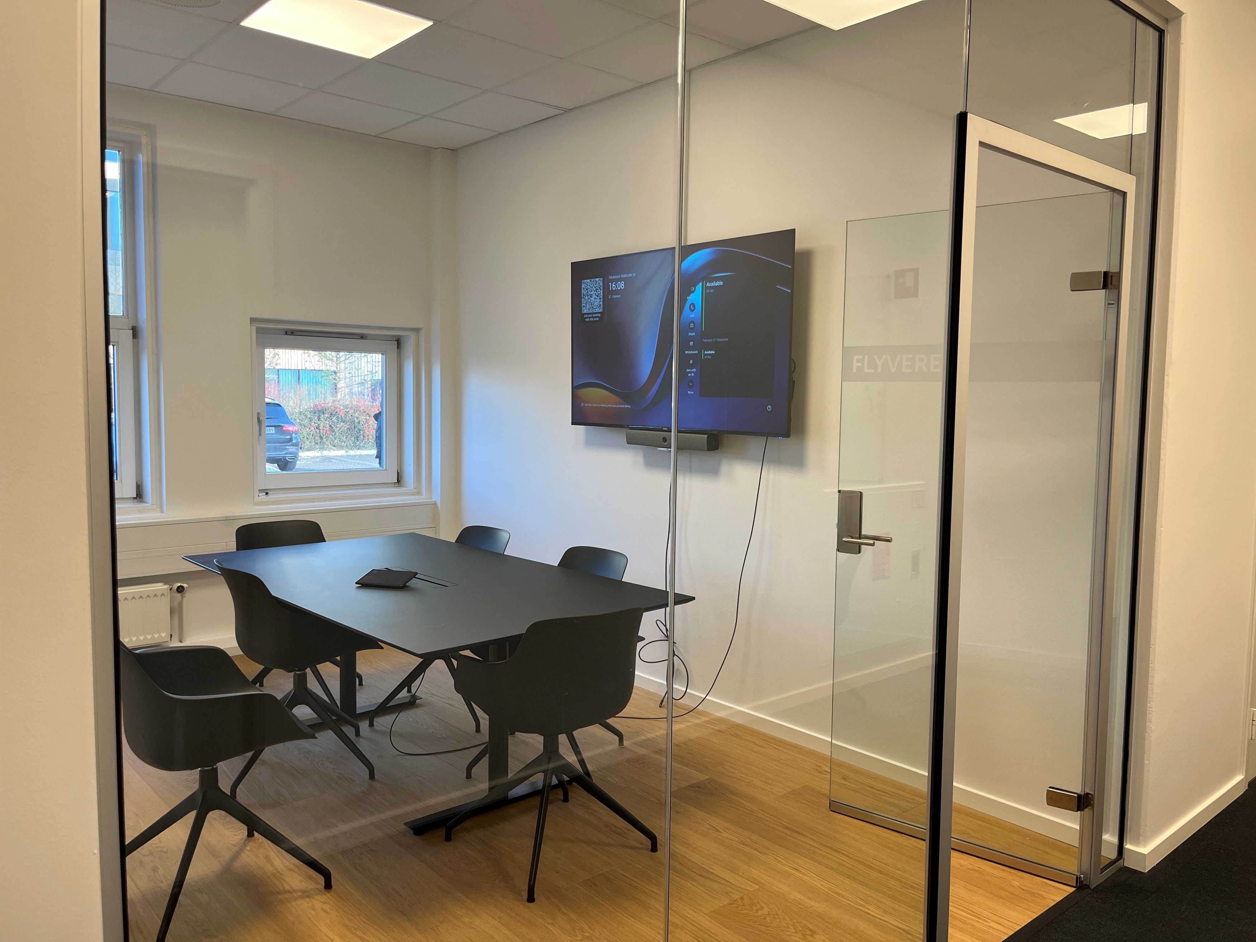 Mødelokale med to store glasdøre, sort bord og stole, monitor og radiator ved siden af vinduet