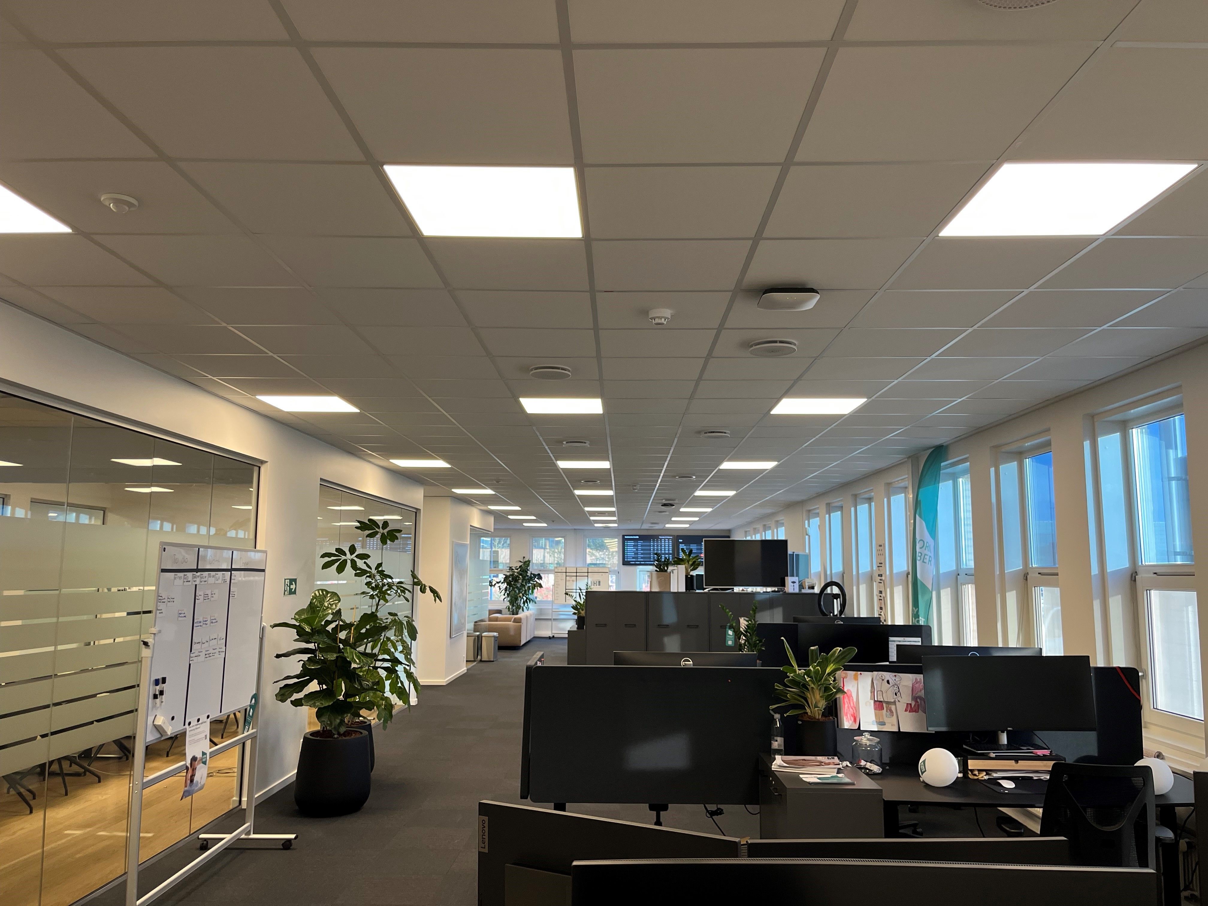 Kontorlokaler med højt til loftet, lyse loftslamper, mange vinduer, glasdøre og lysegulv samt kontormøbler og computere