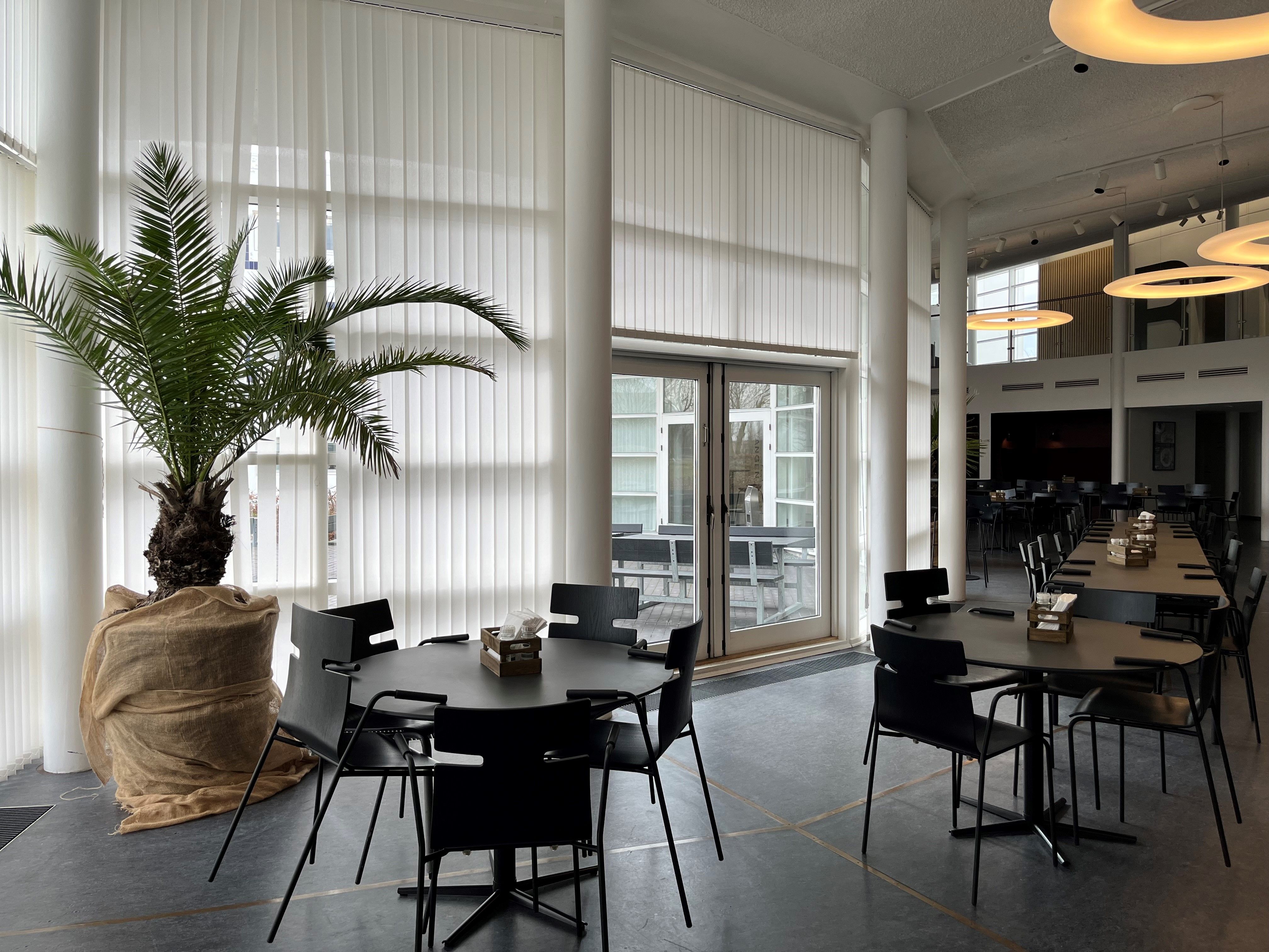 Store hvide jalousier og lyse gulve i en restaurant med runde borde og lyse kroner samt udsigt til gårdhave.