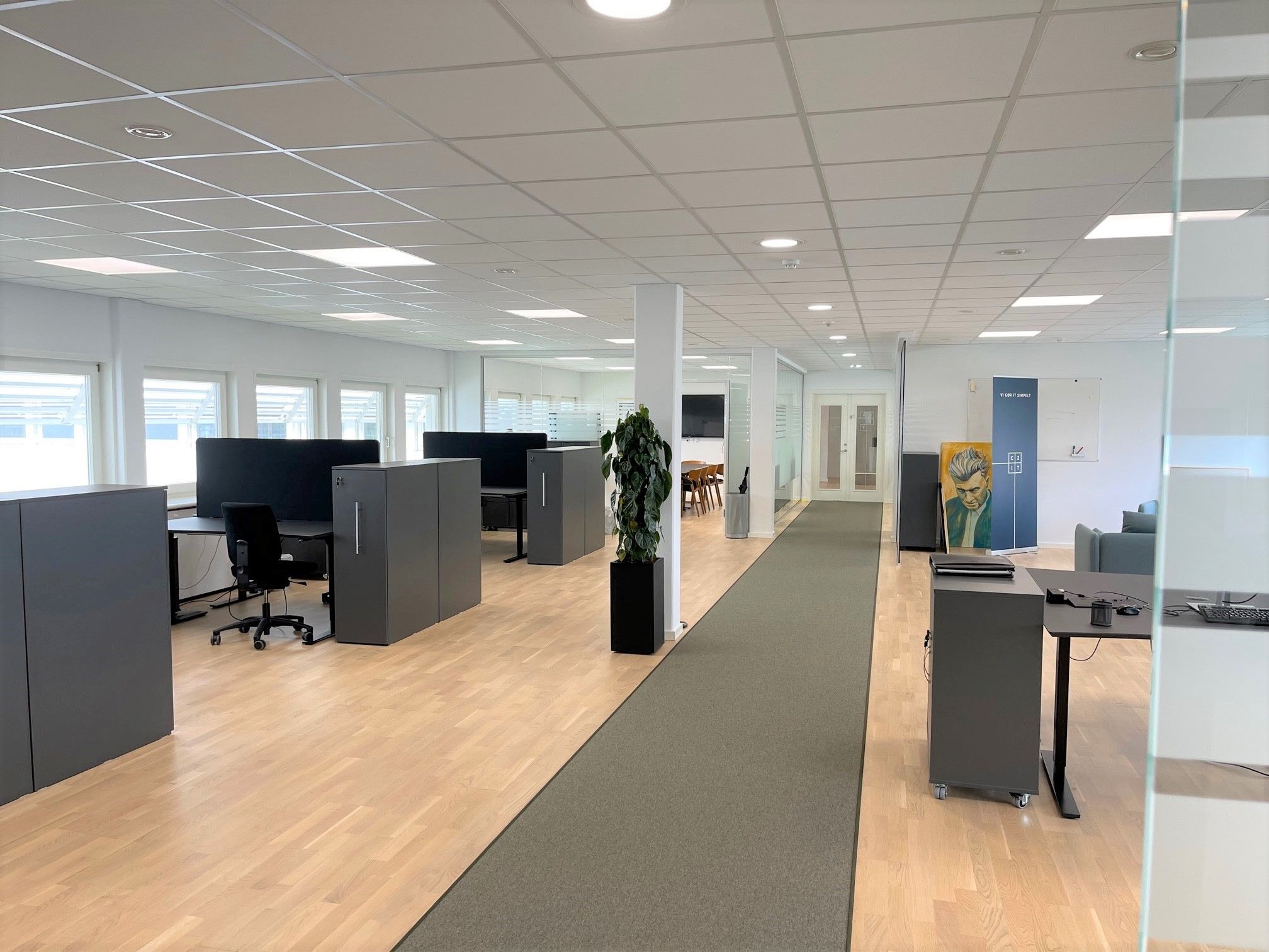 Kontor med møbler og plante i højre side. Vindue i højre side og flere møbler med computere. To bord i højre side og