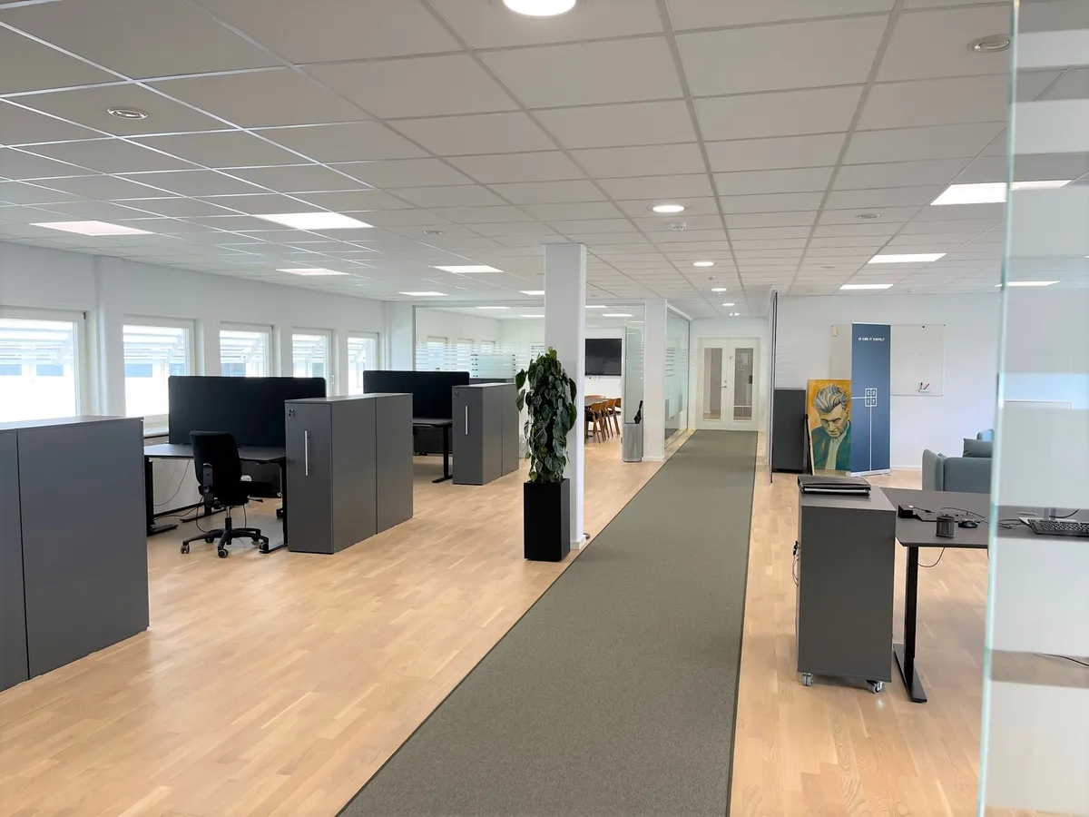 Kontor med møbler og plante i højre side. Vindue i højre side og flere møbler med computere. To bord i højre side og