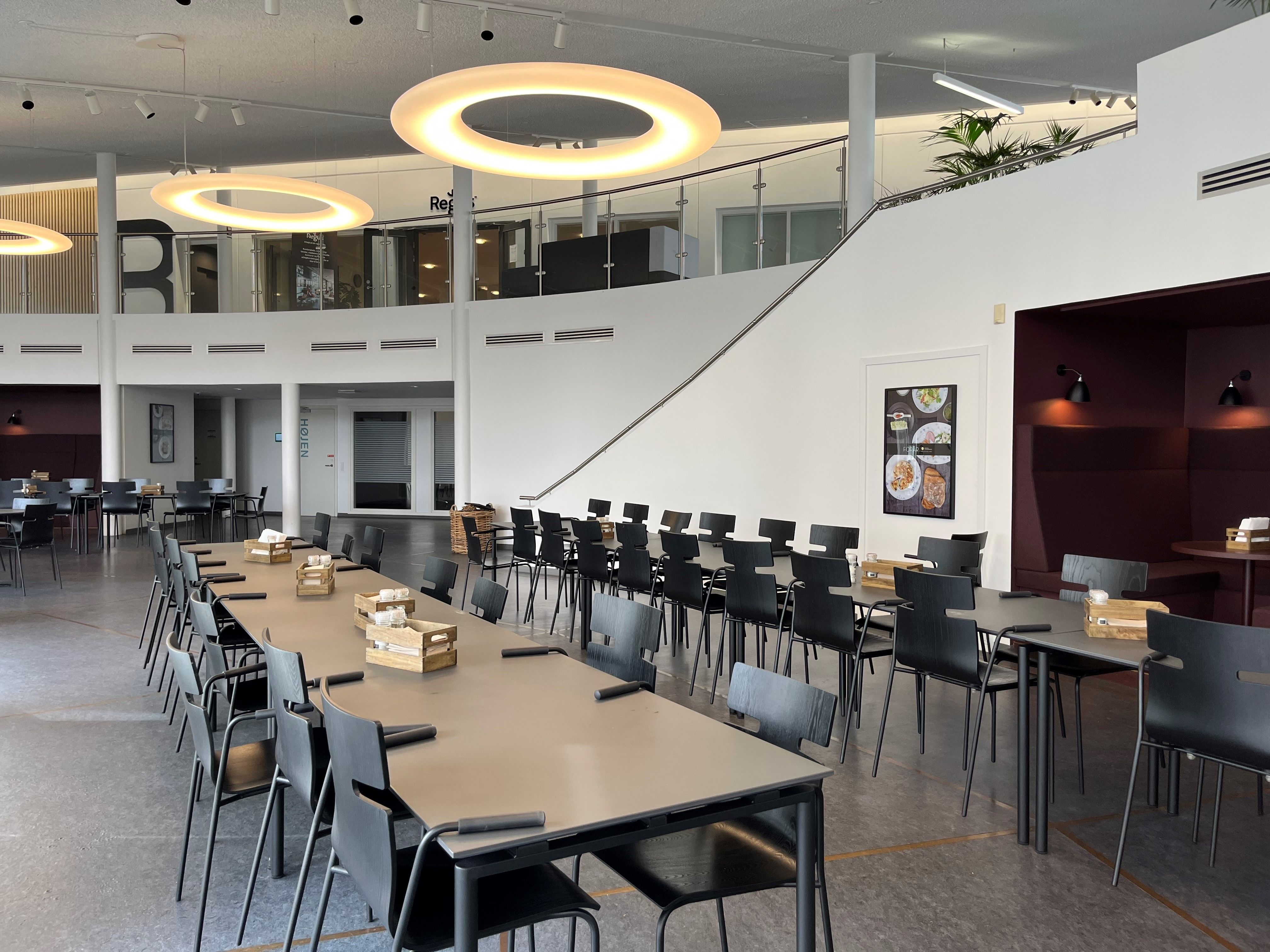 Inde i et lyse, moderne kontor er der lange borde med stole, sammen med et køkkenbord med bønner og et køleskab.