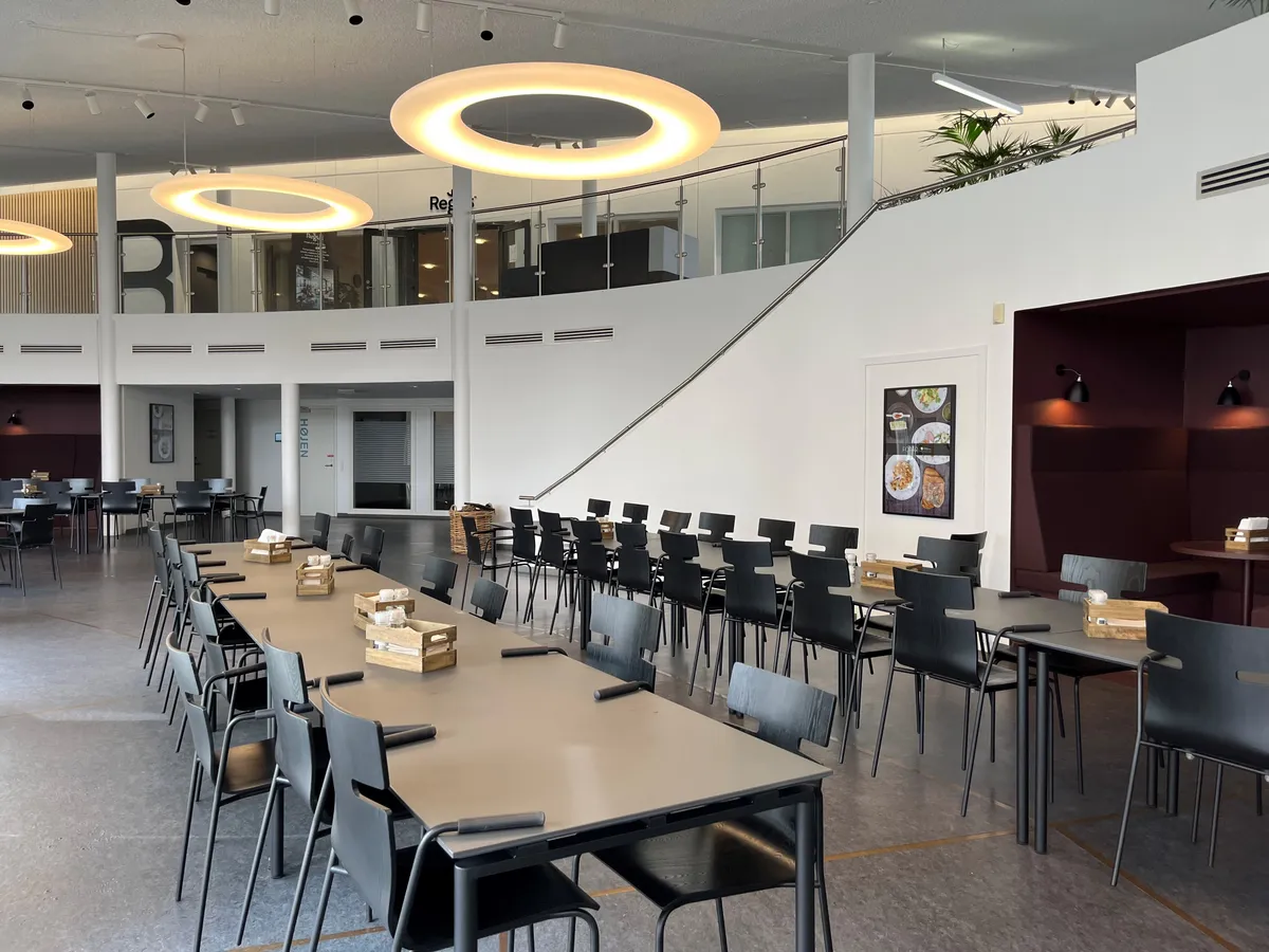 Inde i et lyse, moderne kontor er der lange borde med stole, sammen med et køkkenbord med bønner og et køleskab.