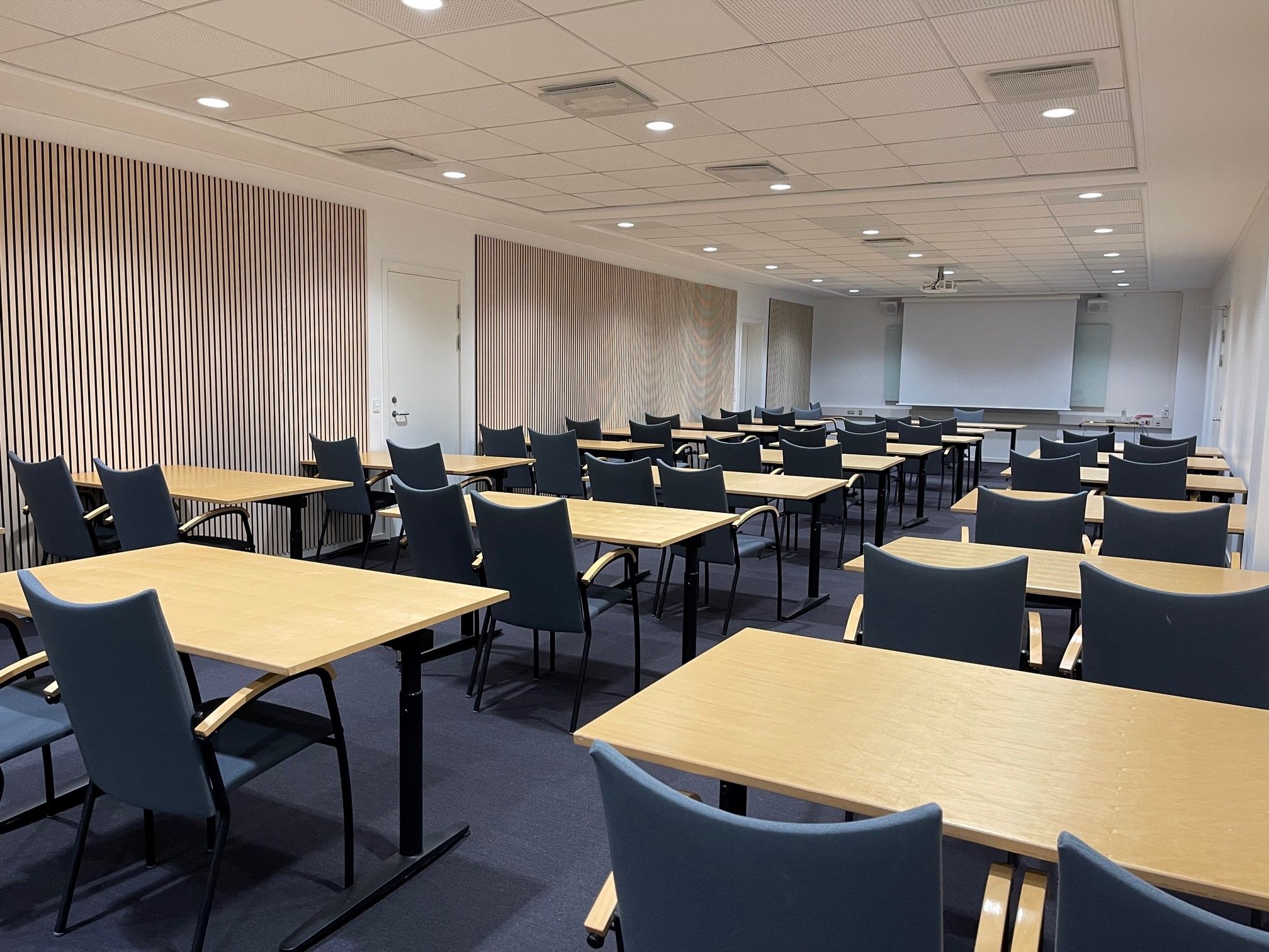 En stor konferencesal med flere borde og stole, moderne møbler, hvide vægge, en hvid projektionsskærm og lygter i loftet