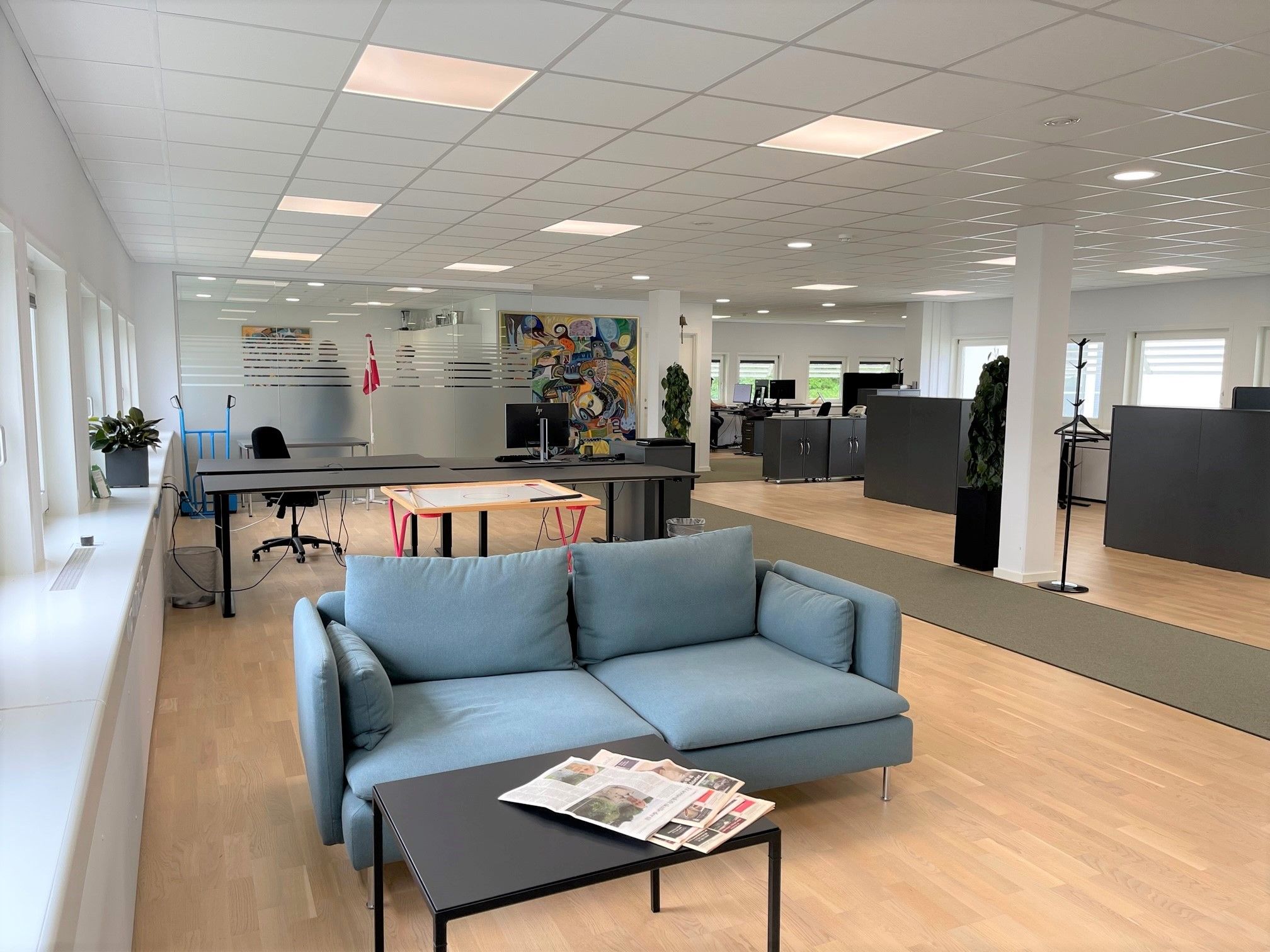 Lejlighed med sofa, bord og aviser, computere og plantepoter, kontorfurniture og kunst på væggen