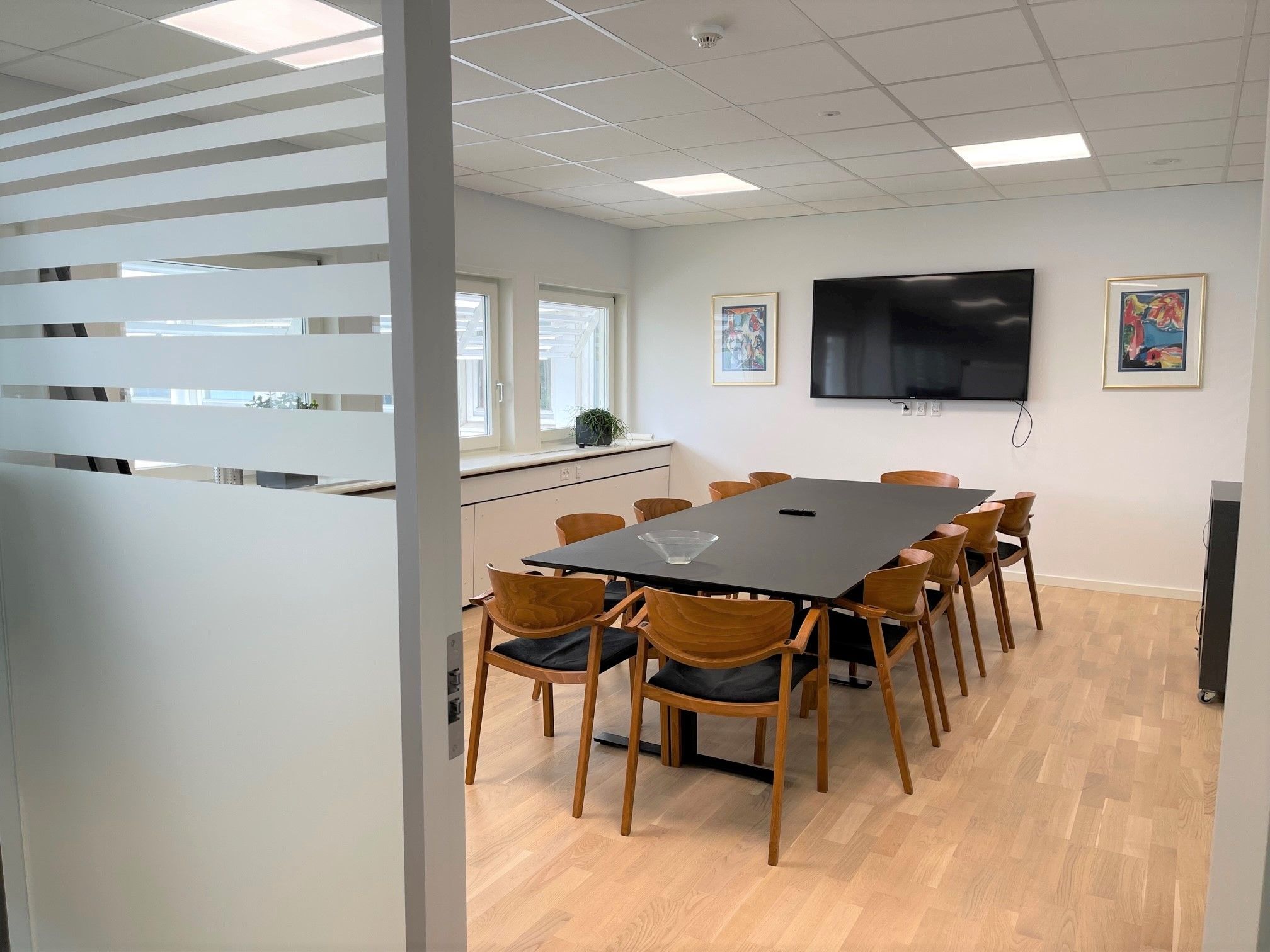 Moderne mødelokale med 12 stole, et skrivebord og en stor tv-skærm. Foran skrivebordet er der et gulvvasket glas og et lille