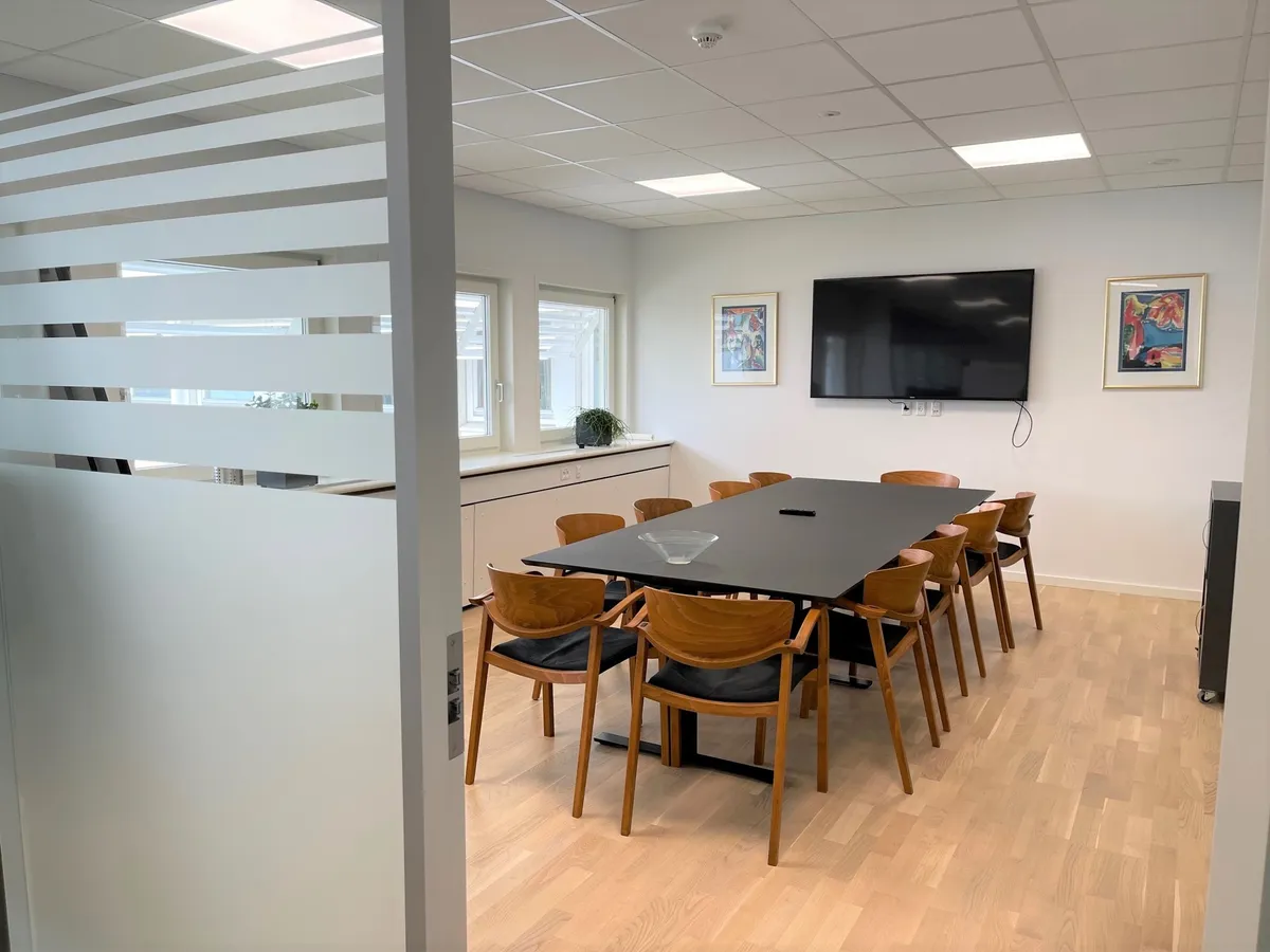 Moderne mødelokale med 12 stole, et skrivebord og en stor tv-skærm. Foran skrivebordet er der et gulvvasket glas og et lille