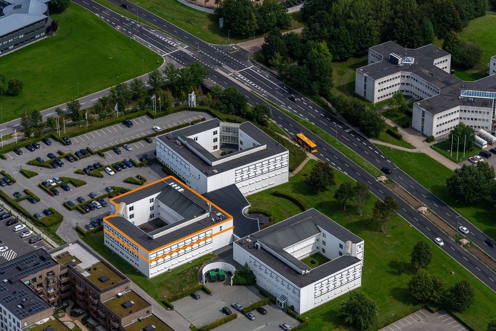 Luftfoto af et stort bygningsanlæg med mange parkeringspladser og en stor hovedvej, der går forbi