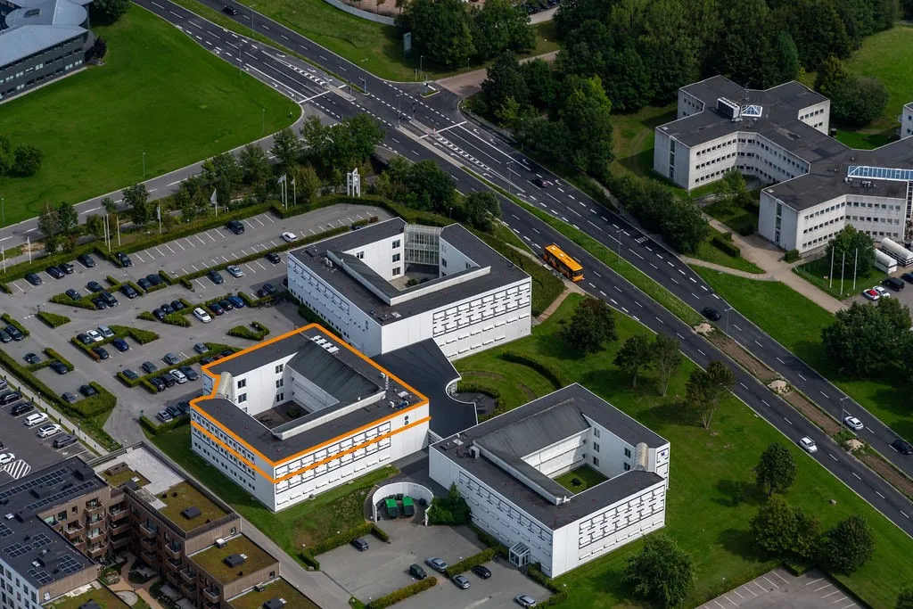 Luftfoto af et stort bygningsanlæg med mange parkeringspladser og en stor hovedvej, der går forbi