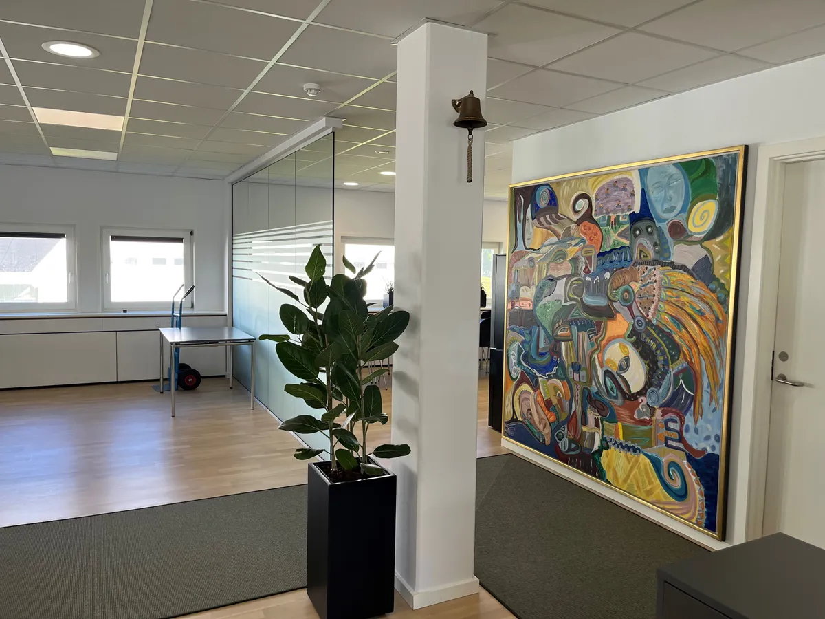 Et stort maleri hænger på væggen. En stor plante står i en potte i midten af rummet. Der er et bord med hjul og en lampe på