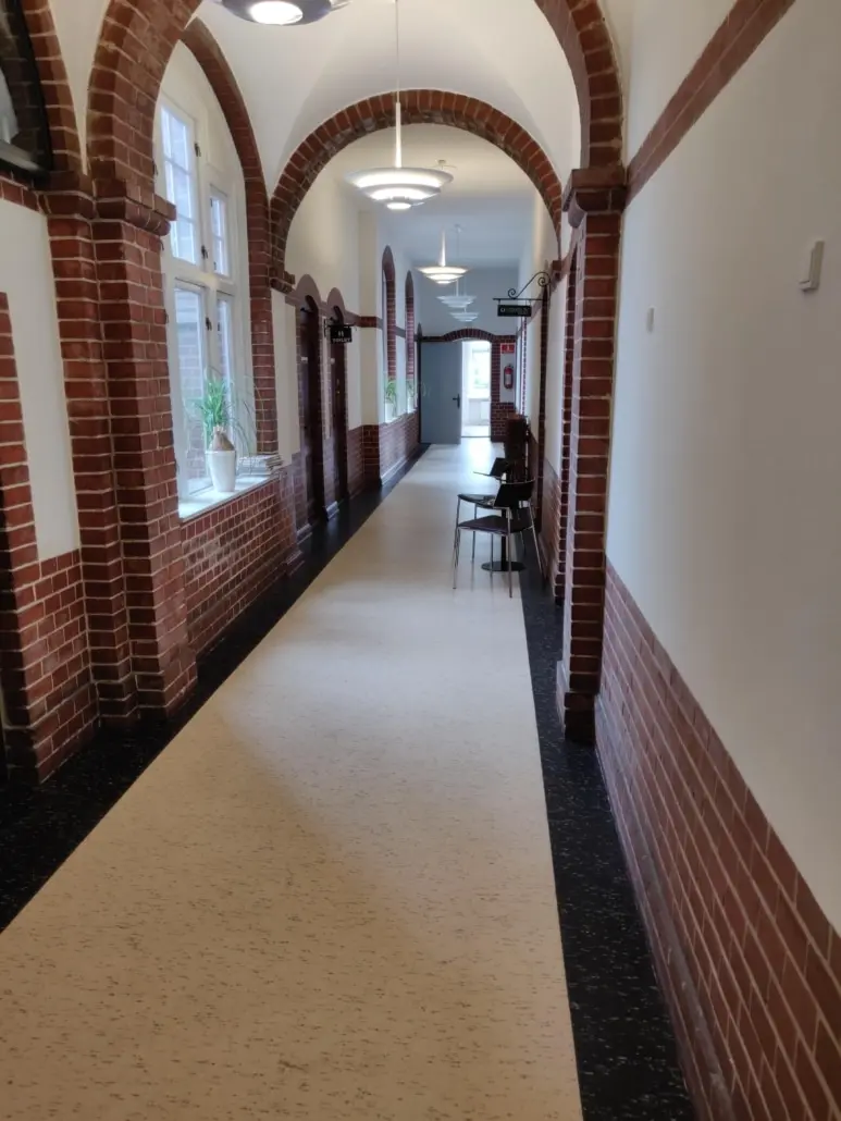 Lang, bred korridor med højbejle og lysekroner, slanke søjler og røde murstensvægge. To stole med bord og et plantepot