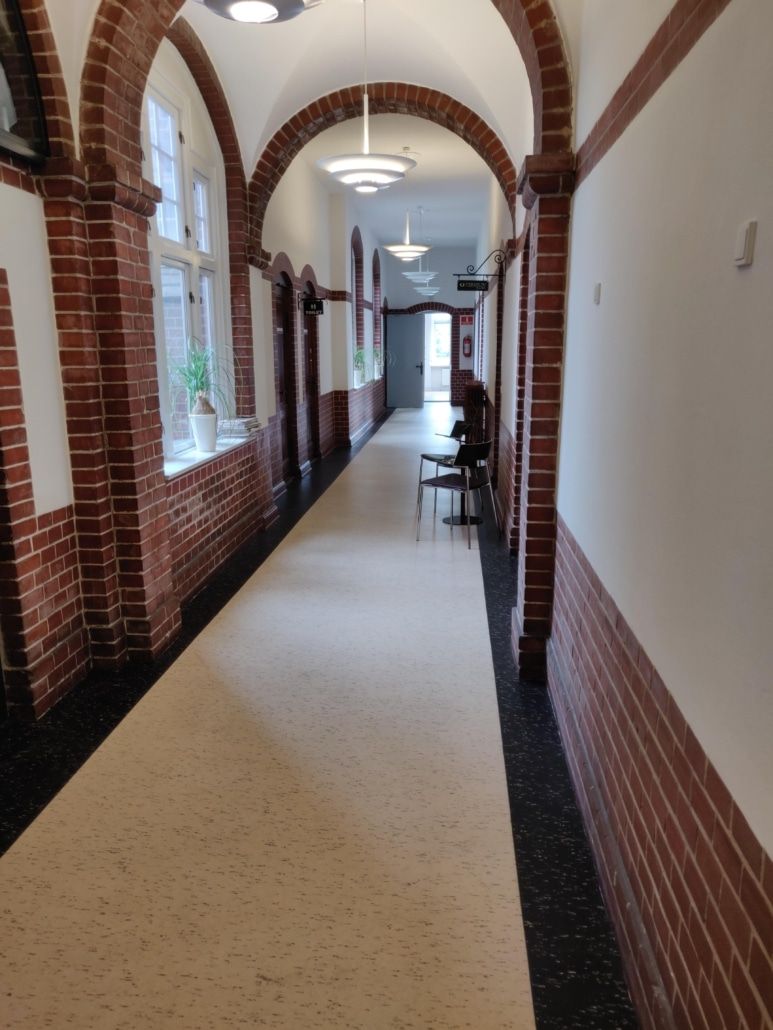 Korridor med højbuede hvælvinger, hvide vægge og røde mursten. Møbler og grønne planter står på vindueskarmen. Hallen er
