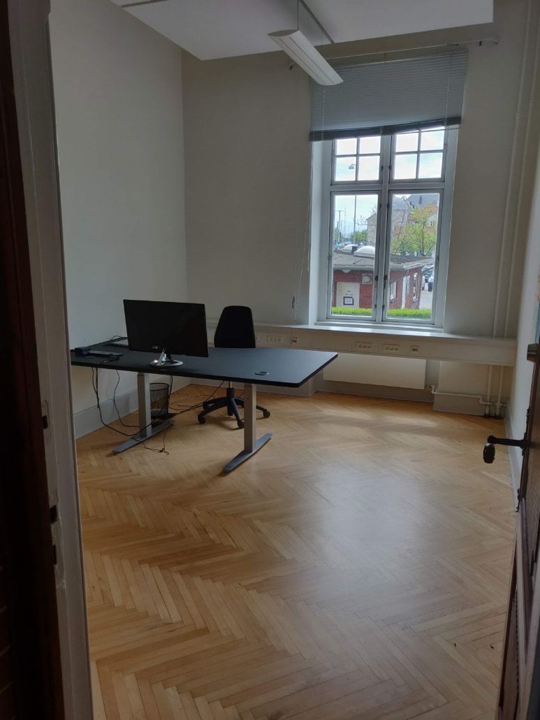 Et moderne kontor med to borde, en stol, to skærme og en radiator under vinduet. Loftet er hvidt og har et stort lys. Gulvet