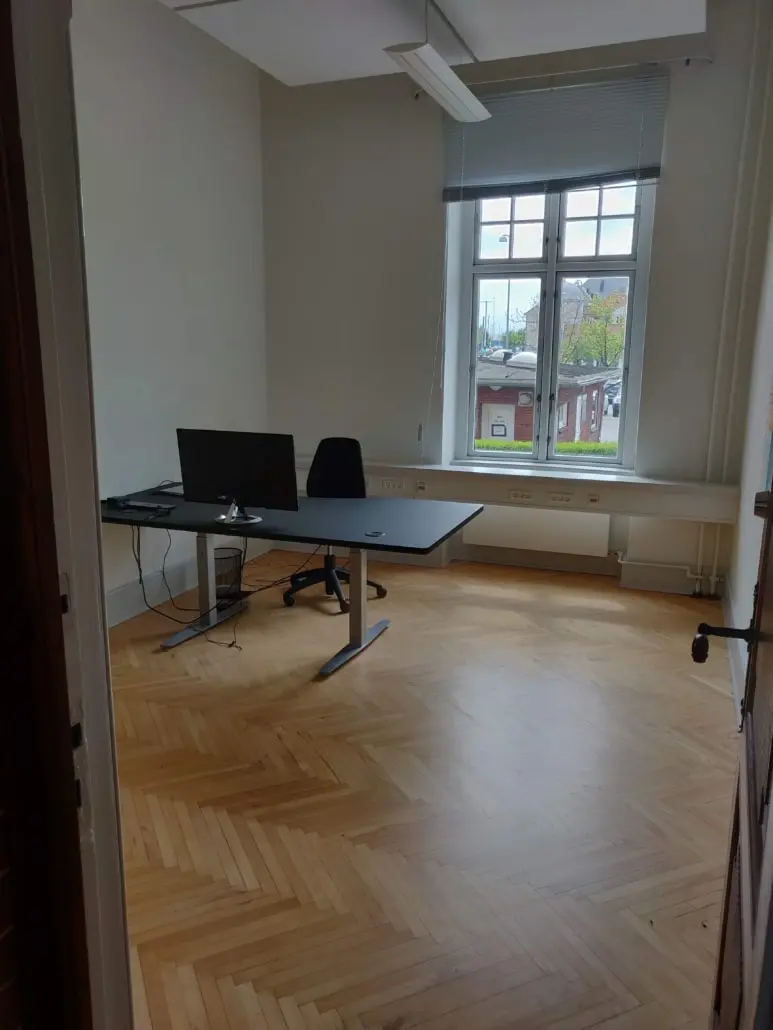 En tom kontor med en skrivebord, to skærme, en stol og en papirkurv. Der er vinduer med jalusi og en hvid gardin. Loftet er