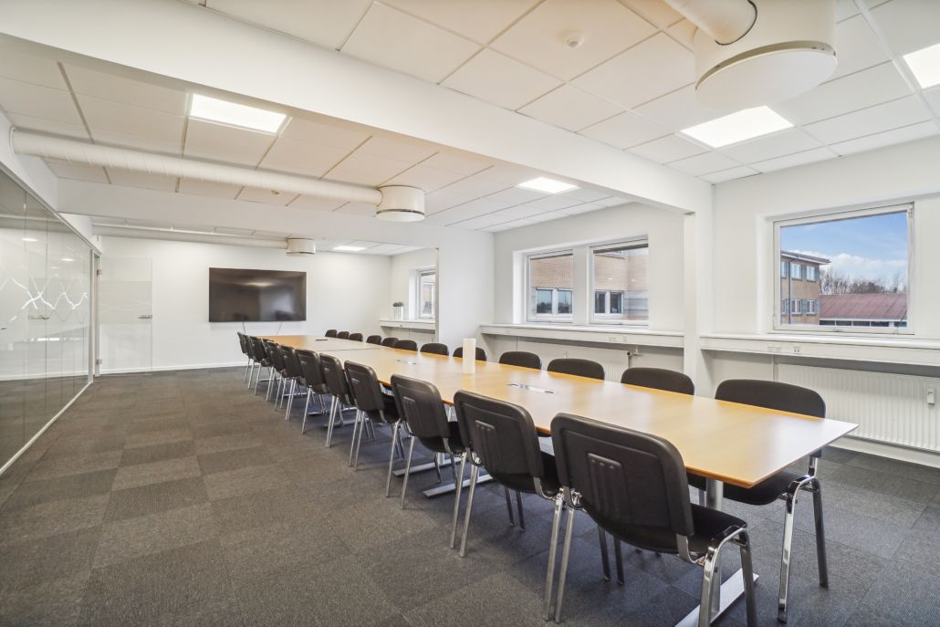 En stor konferencesal med moderne interiør, hvor der er en lang konferencebord med stole og en tv-skærm på væggen