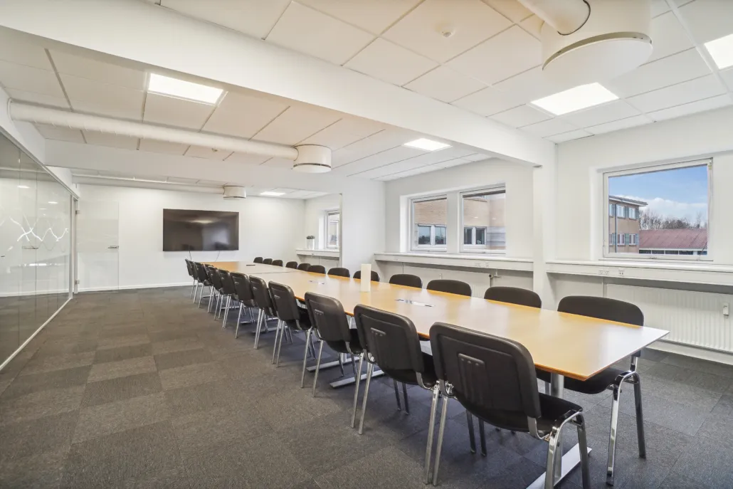 En stor konferencesal med moderne interiør, hvor der er en lang konferencebord med stole og en tv-skærm på væggen