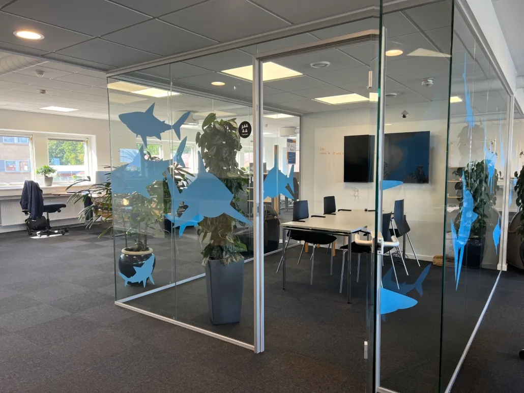 Kontor med moderne indretning og en stor plante i grå potte. Glasskærme med blå haje på. En hvid skrivebord med flere stole.