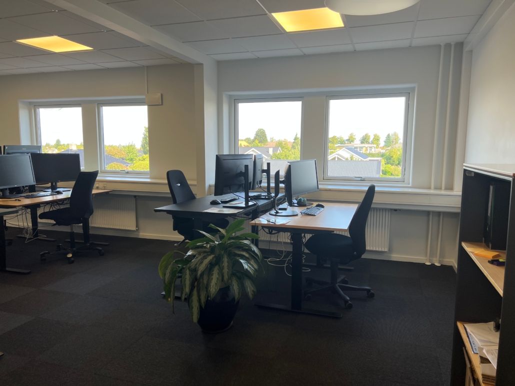 En tom kontor med skrivebord, computer, stol og plante. Væggene er hvide og der er vinduer med udsigt til en park.