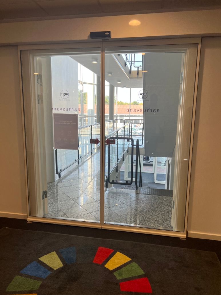 Indegang til Voxeværket i Aarhus med glasdør og stående skrift. Det er et moderne kontor med lysegule vægge og en stor hal