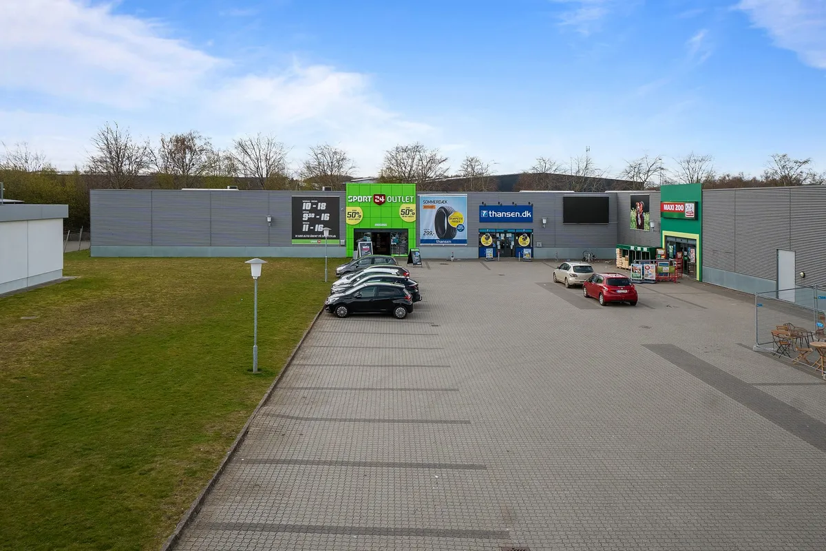 To biler parkeret i parkeringsplads med mærker Sport 24 Outlet og MAXI 200, flere butikker og grønne arealer i baggrunden